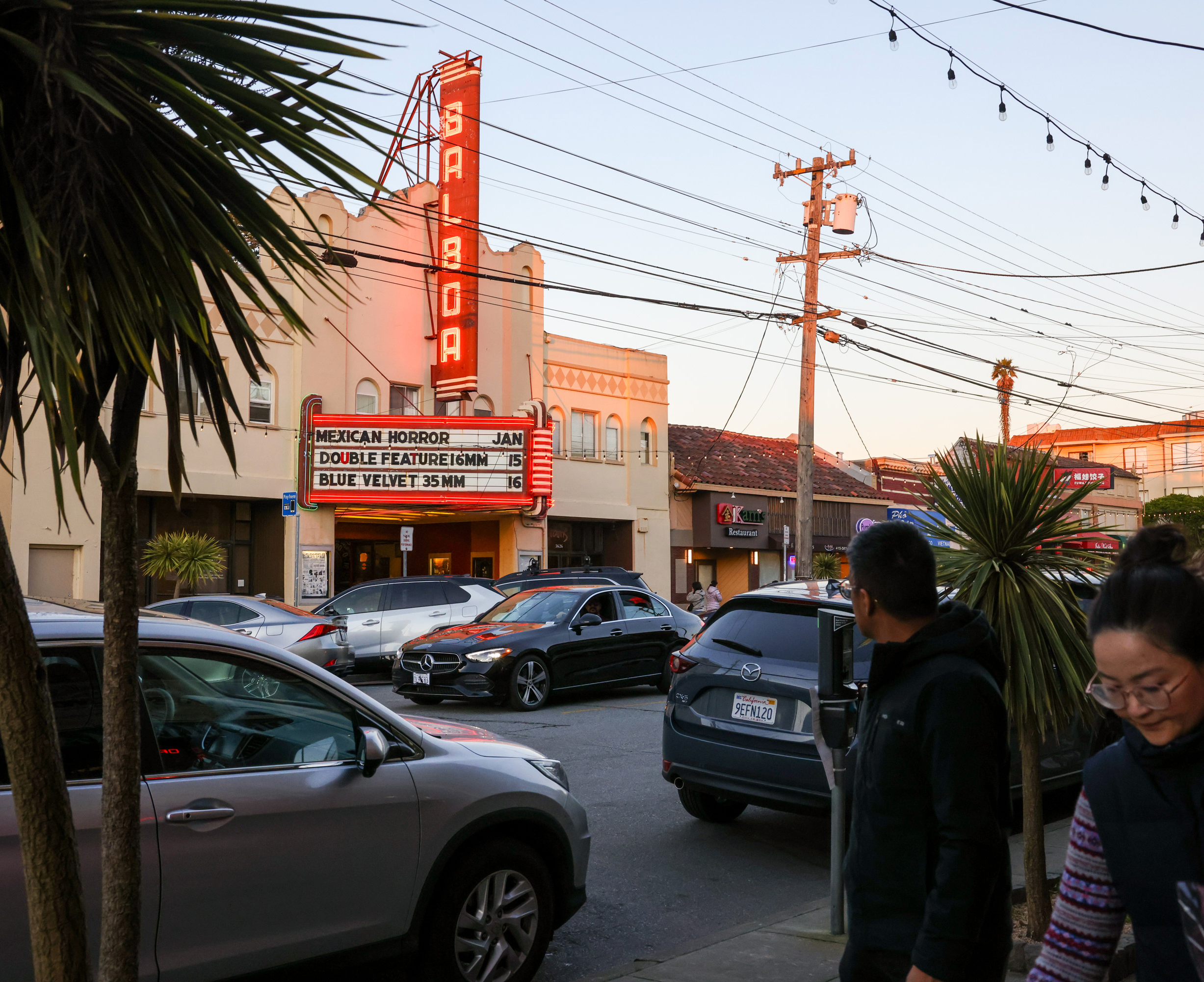 A street scene with a theater marquee showing movie titles, cars parked along the road, and two people walking on the sidewalk under string lights.