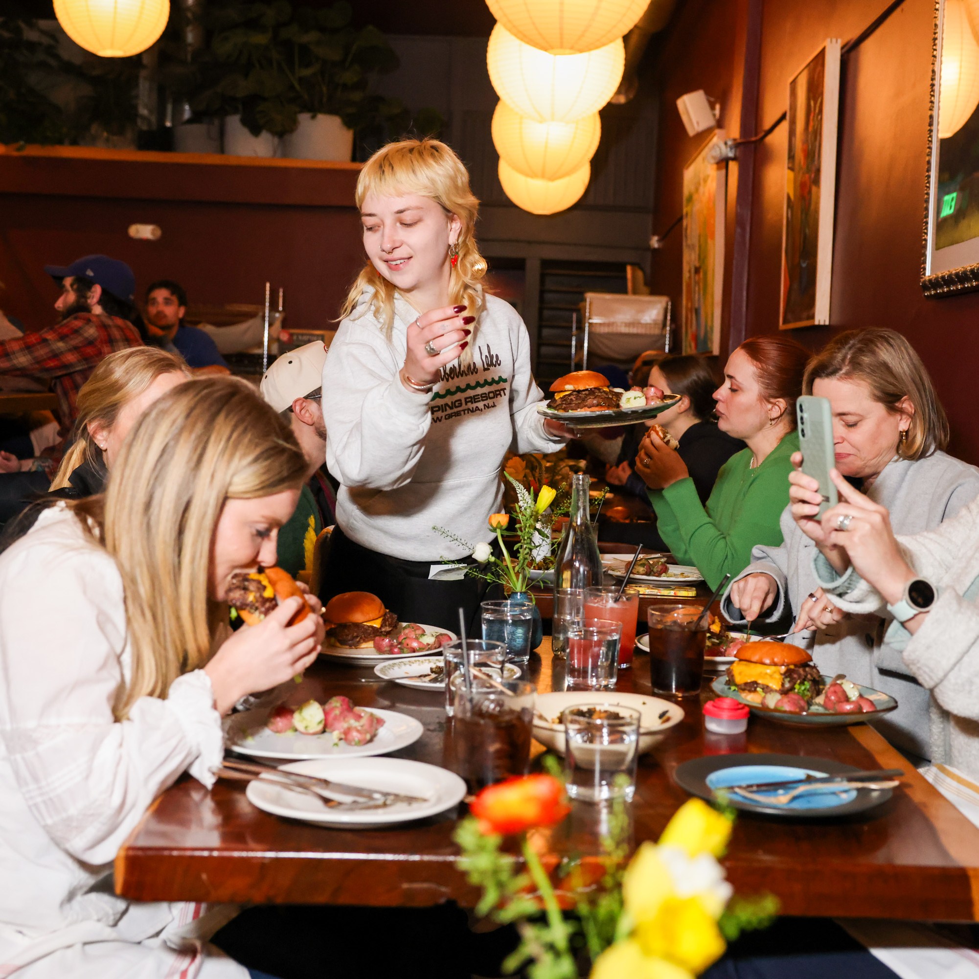 A cozy restaurant scene with people enjoying burgers and drinks. A person in white is serving food, while others take photos and chat around a wooden table.