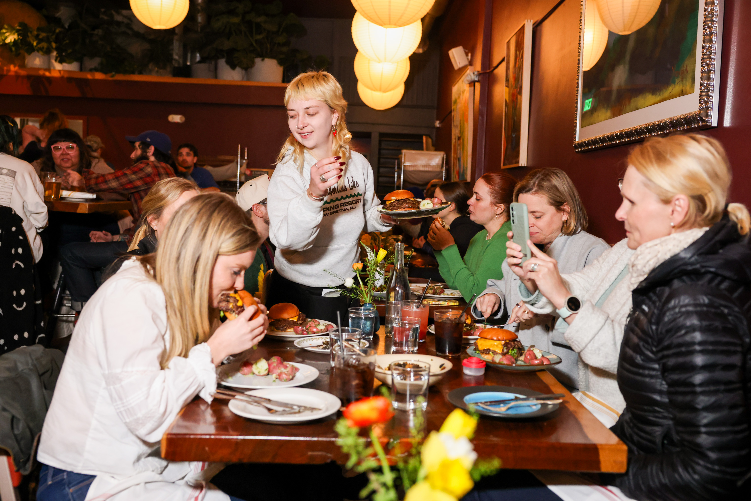 A cozy restaurant scene with people enjoying burgers and drinks. A person in white is serving food, while others take photos and chat around a wooden table.