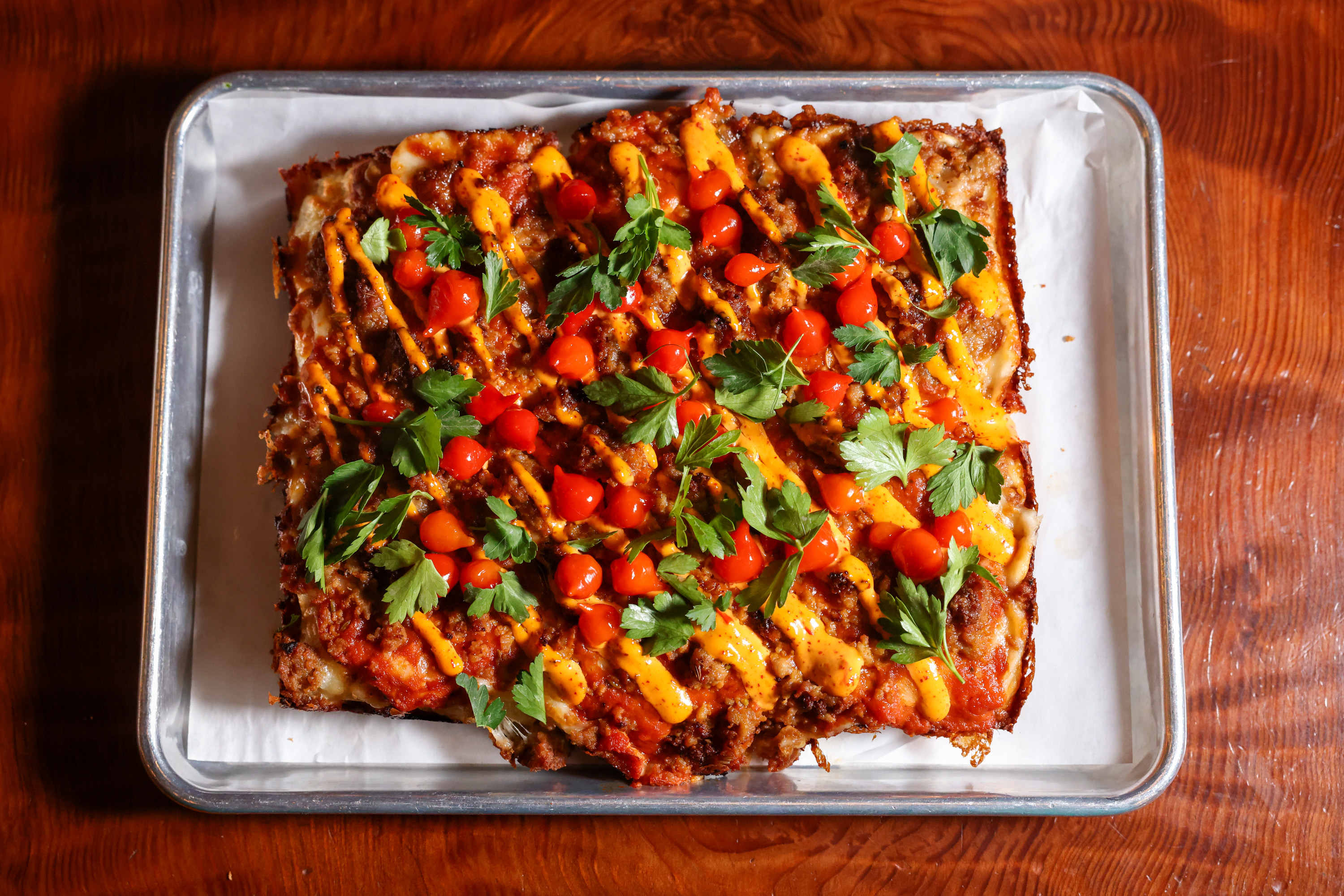 A square pizza on a metal tray is topped with vibrant cherry tomatoes, green parsley, and drizzled with orange sauce on a wooden table.