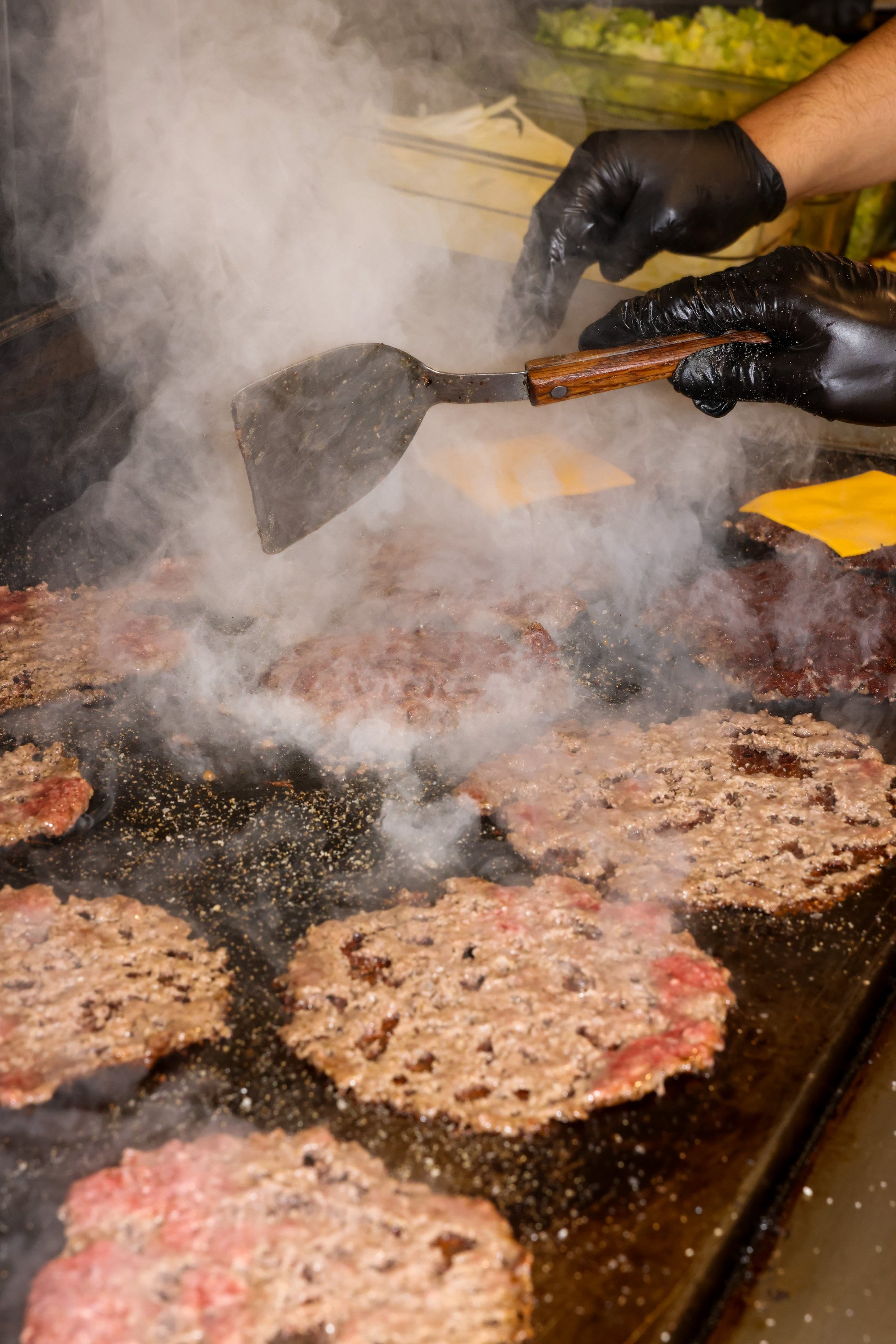 A person wearing black gloves uses a spatula to cook burger patties on a smoky grill, with cheese slices and lettuce visible in the background.