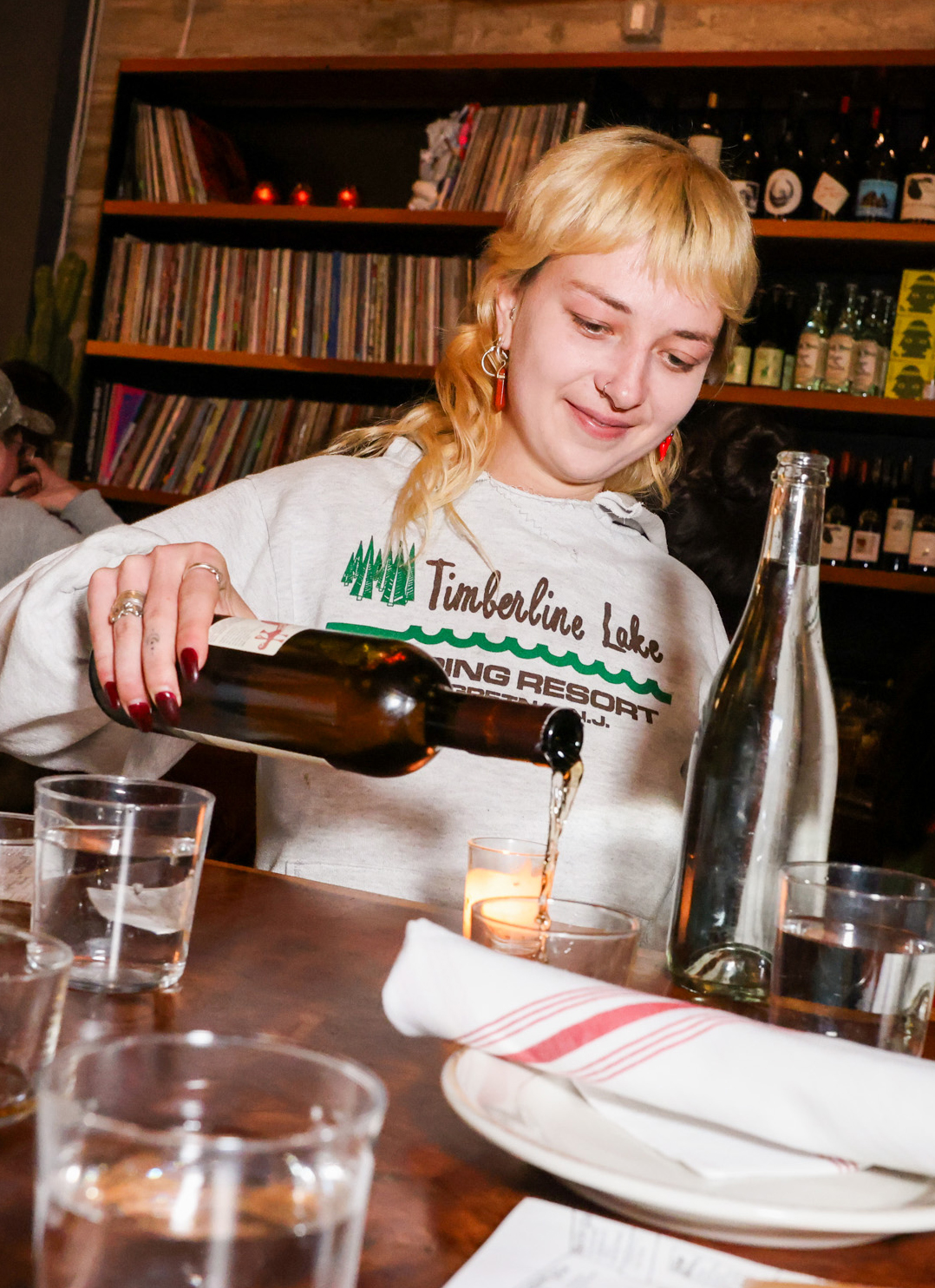 A person with blond hair is smiling while pouring a drink from a bottle into a glass at a table. Shelves with records and bottles are in the background.