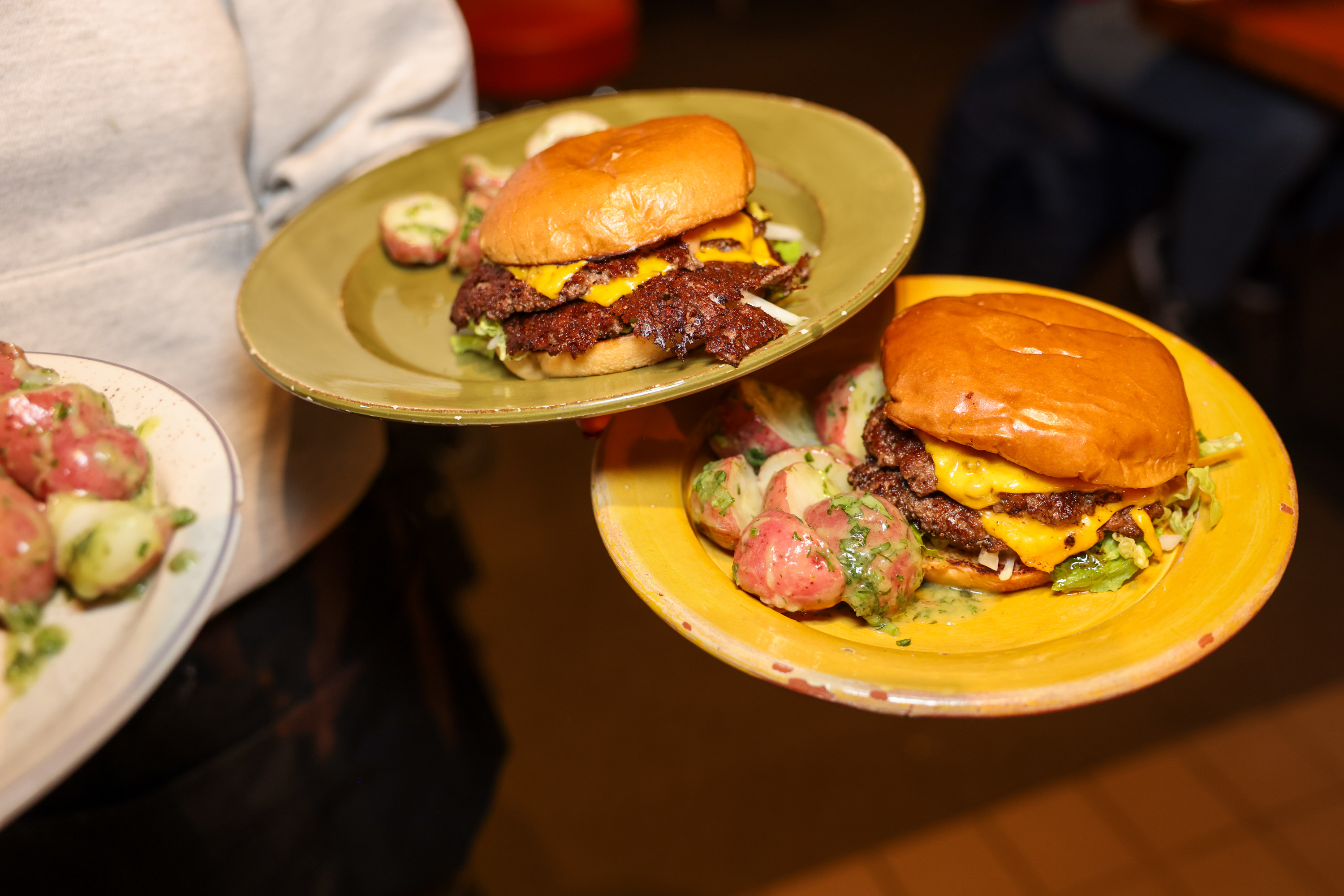 Two plates with cheeseburgers and sides of creamy potato salad are being held by a person in a gray shirt.