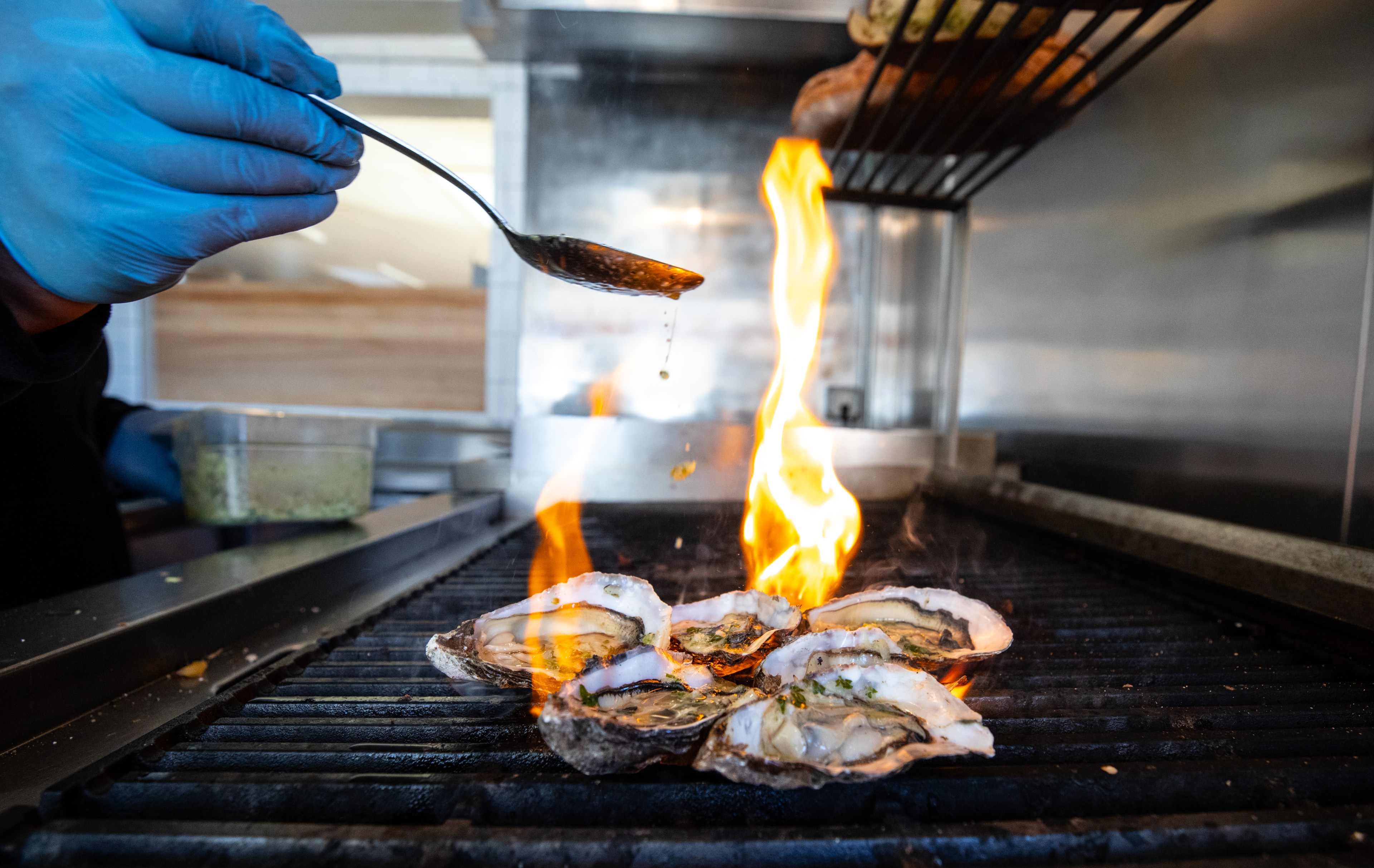 A hand in a blue glove drizzles liquid over oysters on a grill, with flames rising up. In the background, there's a container of herbs or seasoning.