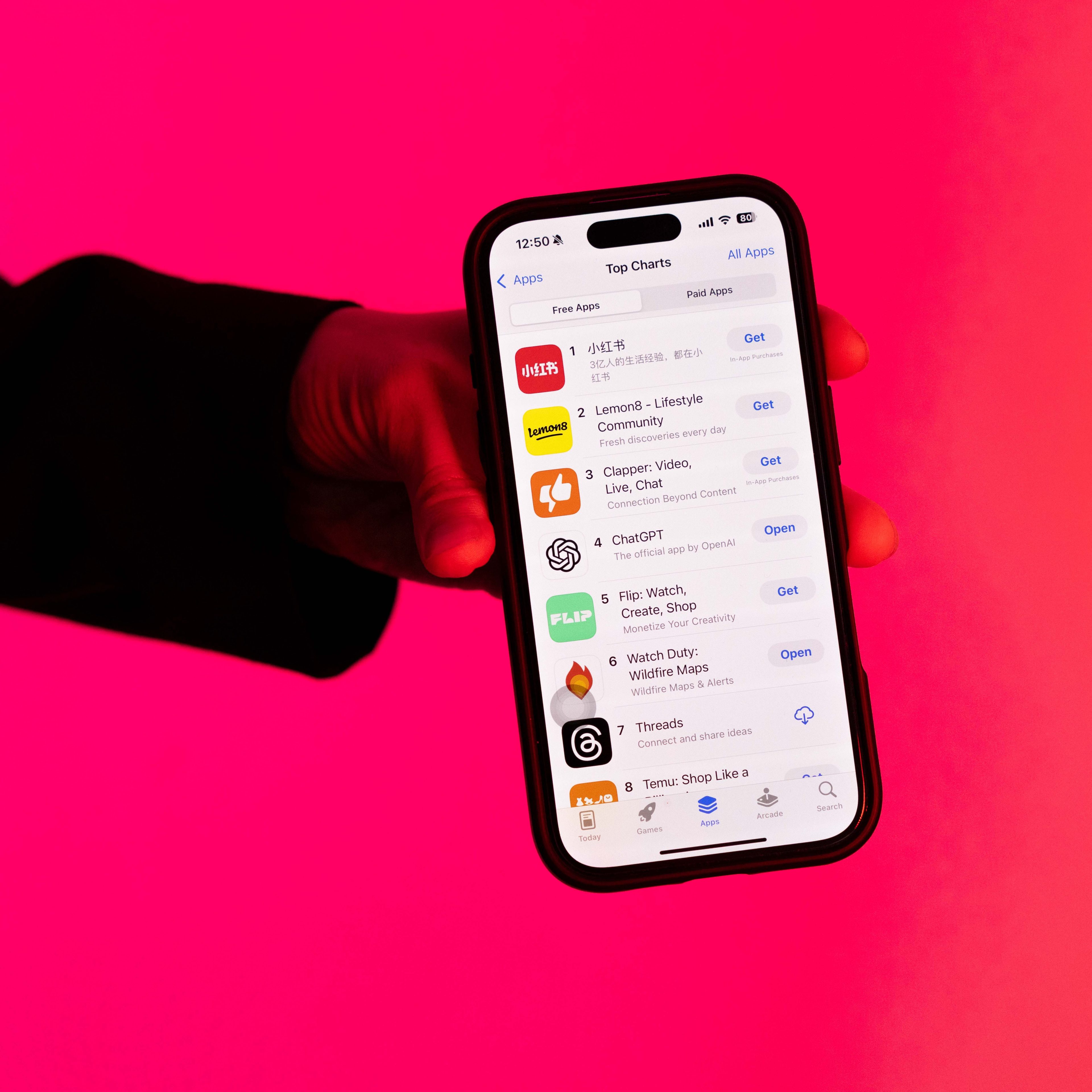 A hand holds a smartphone displaying the App Store's top charts against a bright pink background. Several app icons and names like &quot;ChatGPT&quot; are visible on the screen.