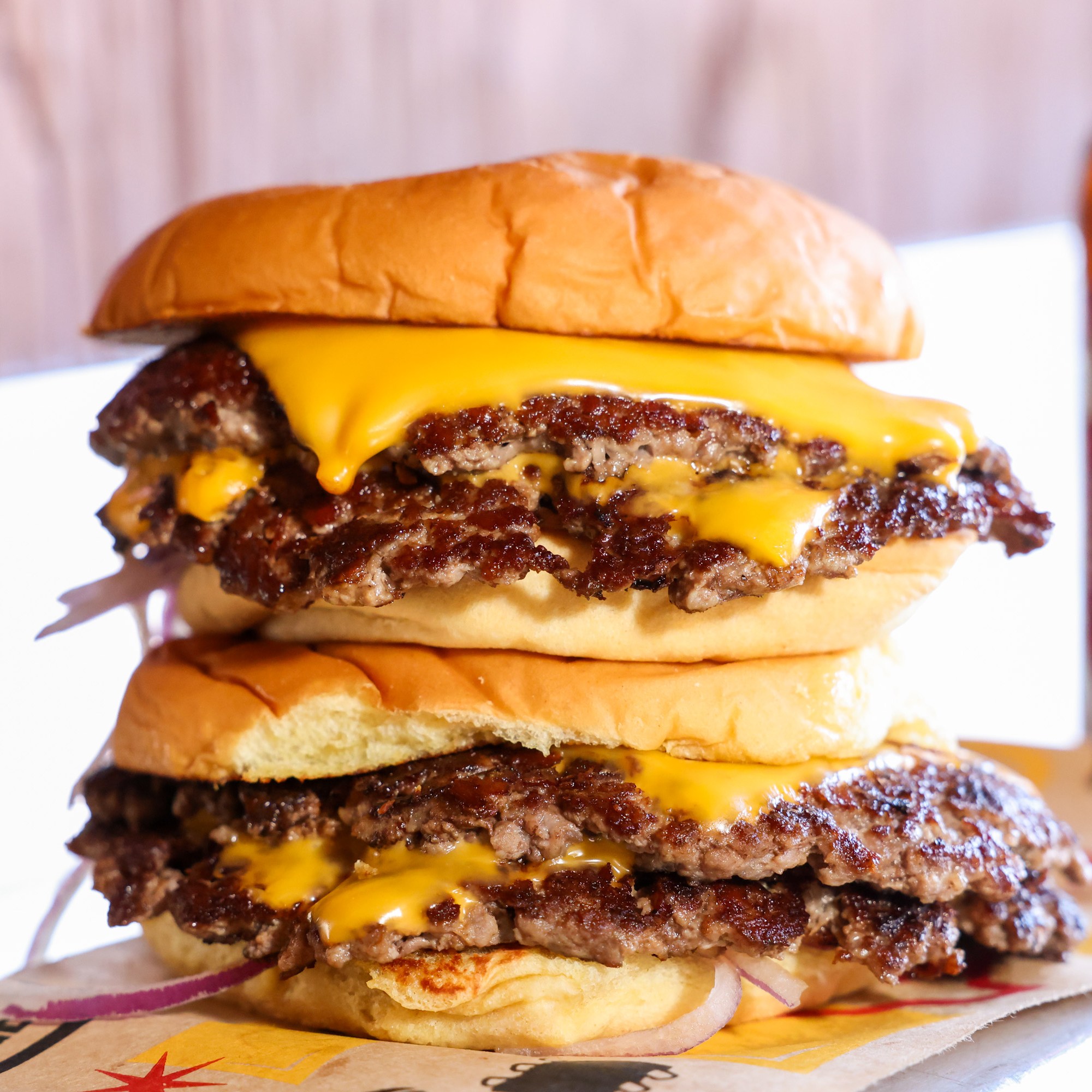 A double cheeseburger with melted cheese and beef patties is stacked high, accompanied by bottles of Coke and beer in the background.