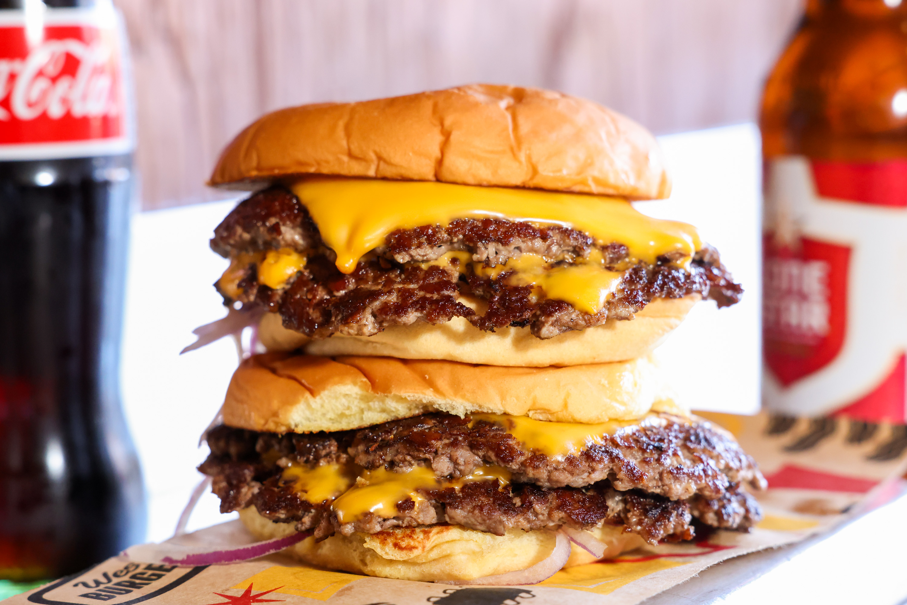 A double cheeseburger with melted cheese and beef patties is stacked high, accompanied by bottles of Coke and beer in the background.