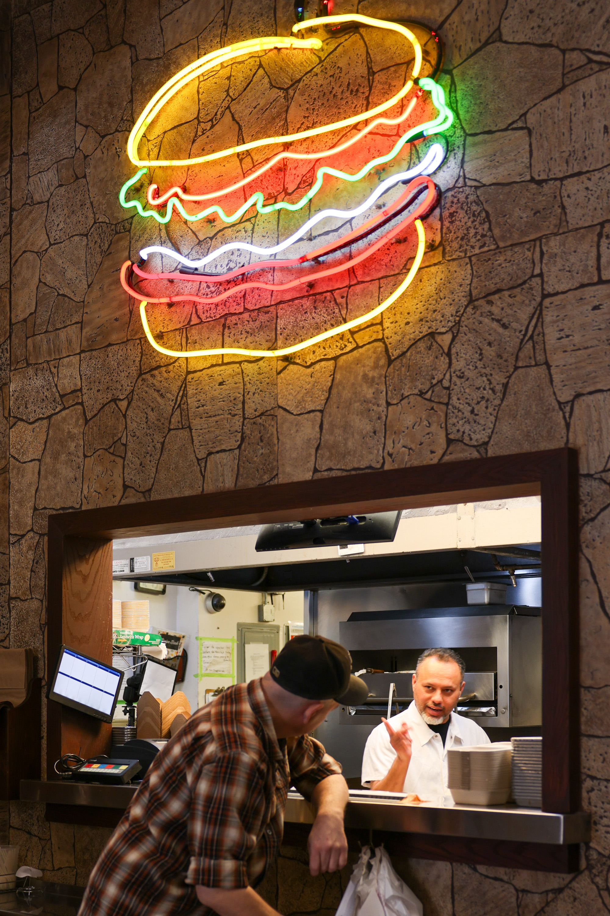 A neon burger sign glows on a stone-patterned wall above a restaurant counter, where a chef speaks with a customer leaning forward in a plaid shirt and cap.