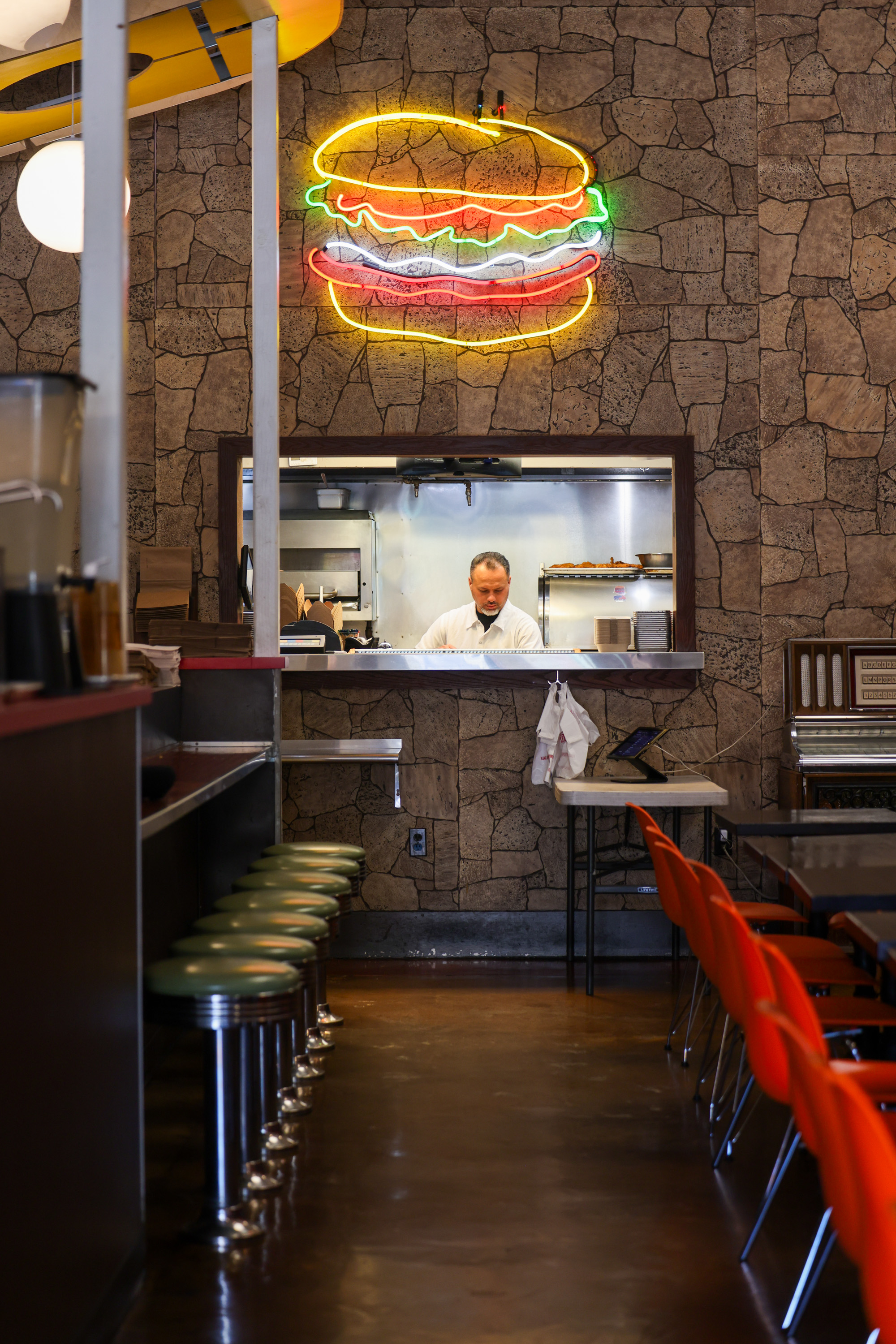 A diner with a stone wall features a neon burger sign. A counter with green stools lines one side, and a man is seen working in the kitchen window.