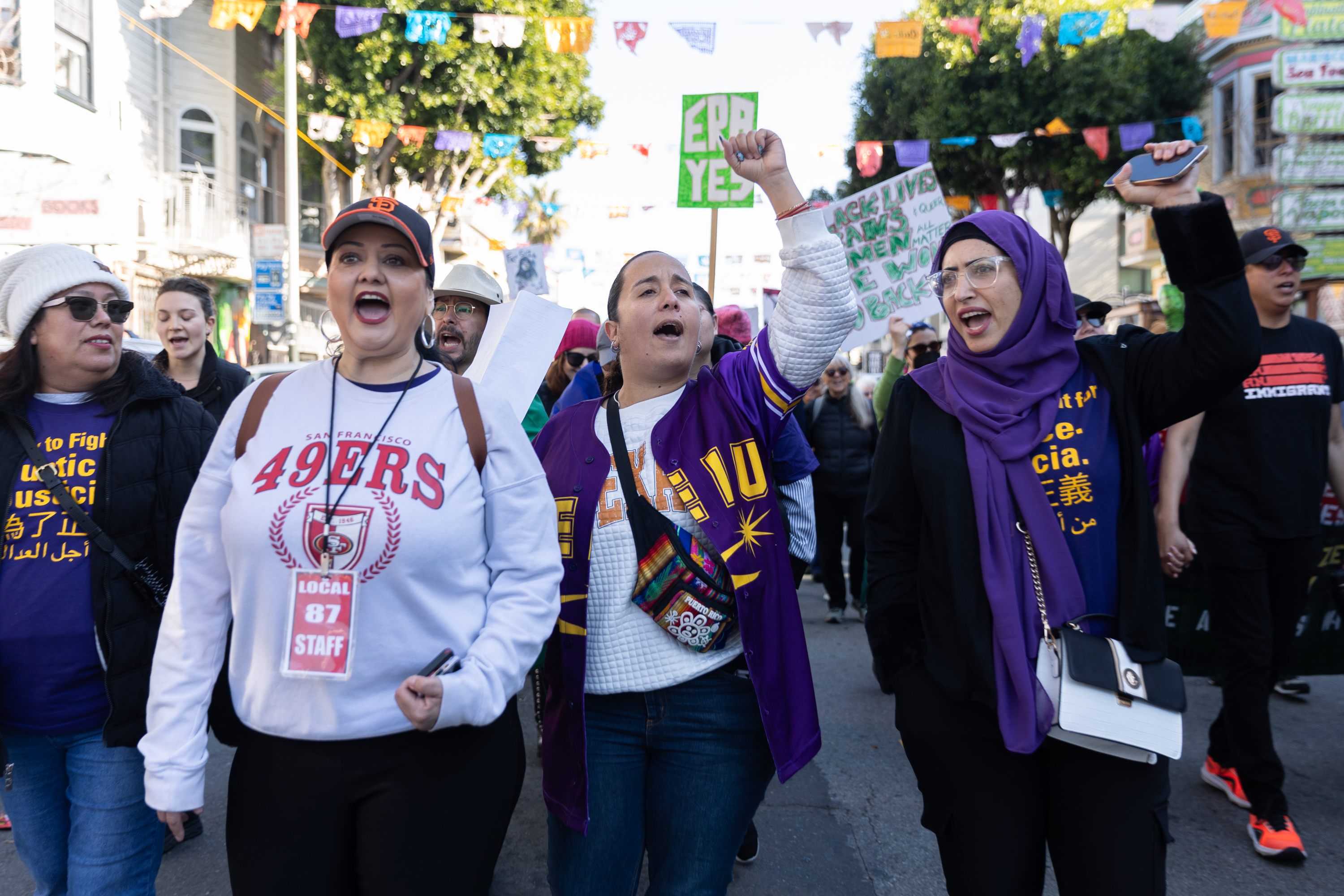 A group of people are marching energetically in a street protest. They wear colorful attire, with one holding a sign that reads &quot;ERA YES,&quot; and others chanting spiritedly.