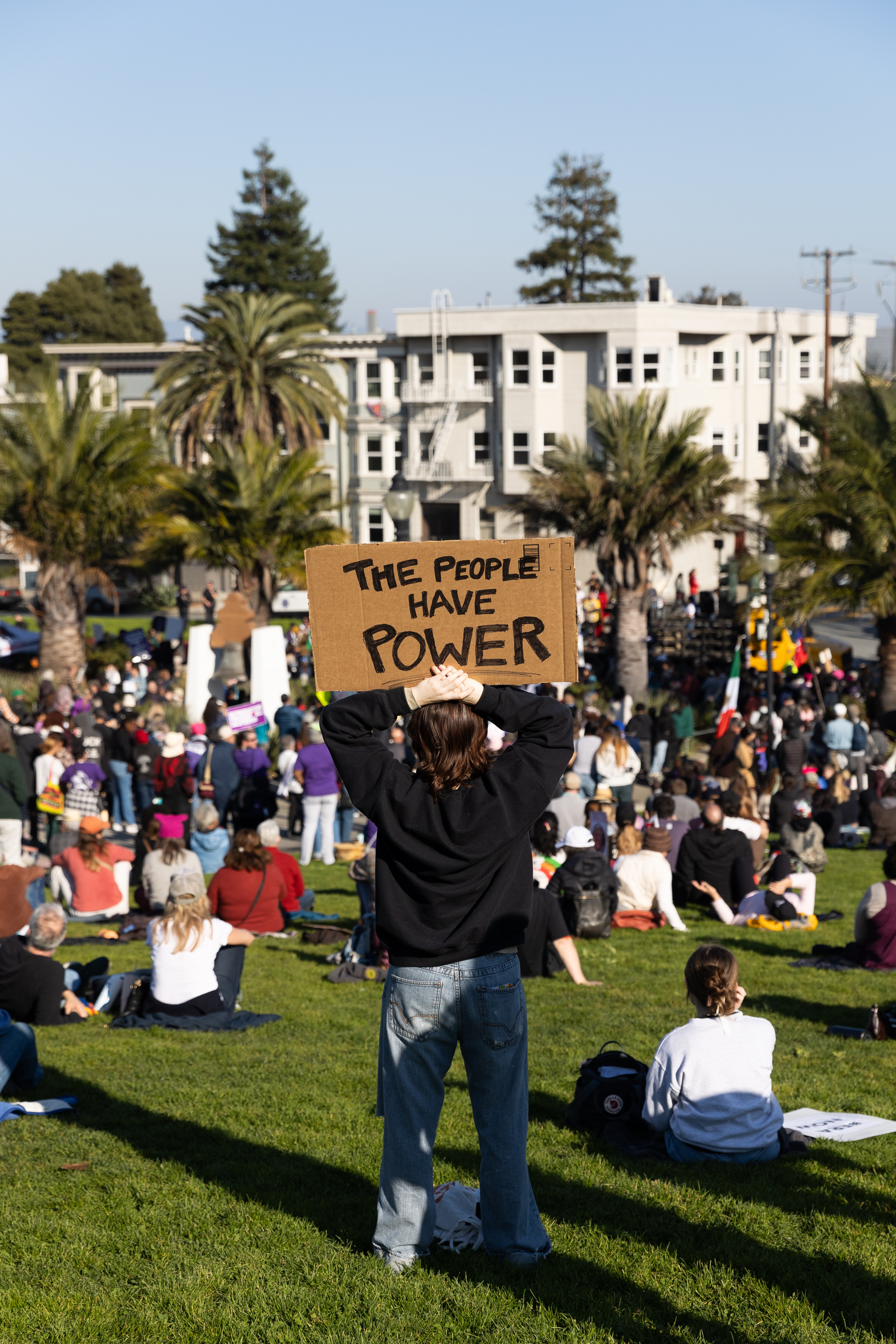 A person holds a sign reading &quot;The People Have Power&quot; amid a crowd at an outdoor gathering, with palm trees and apartment buildings in the background.