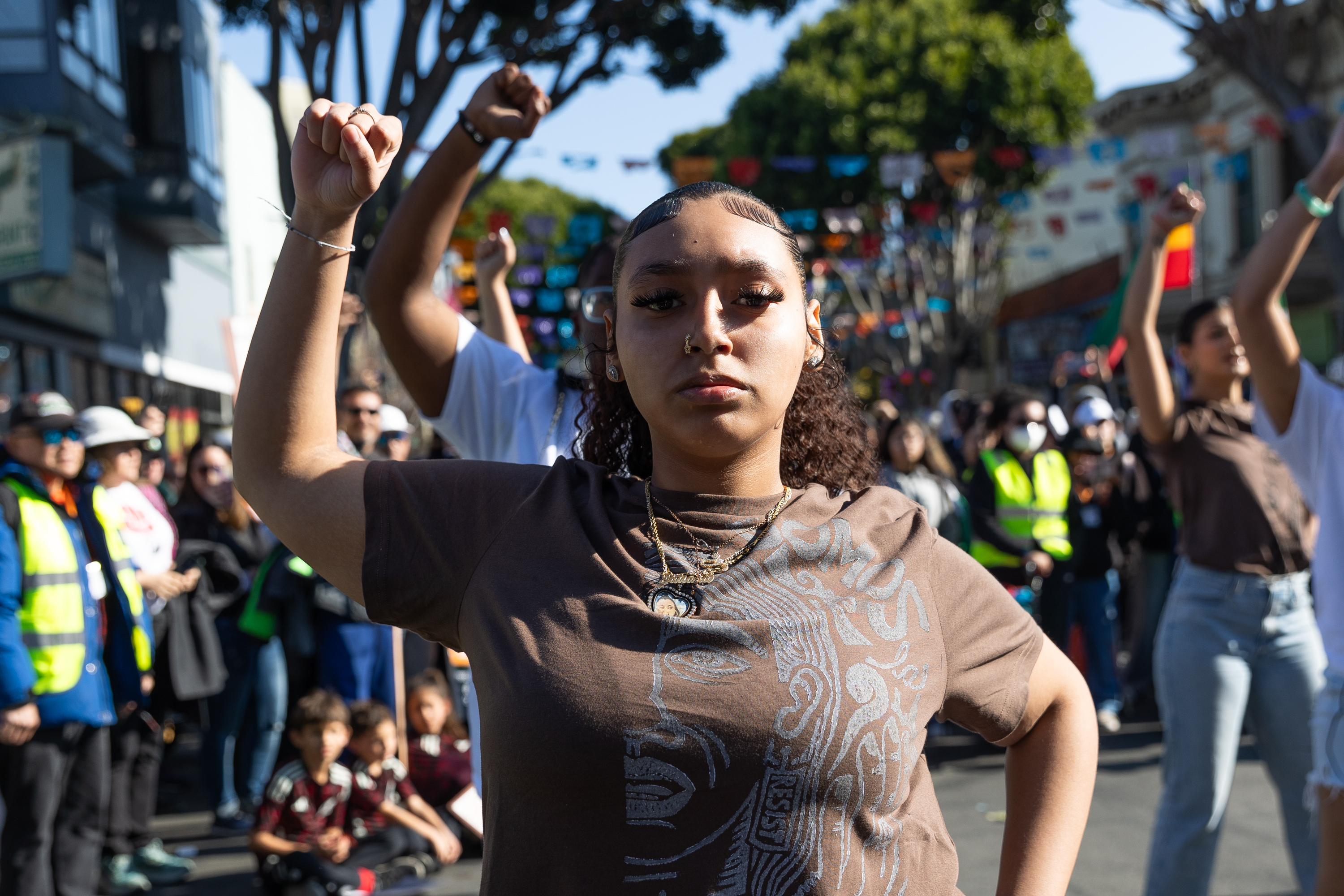 A young woman stands with her fist raised, among a crowd of people outdoors. She wears a brown shirt with a graphic design. Colorful flags and trees are visible in the background.