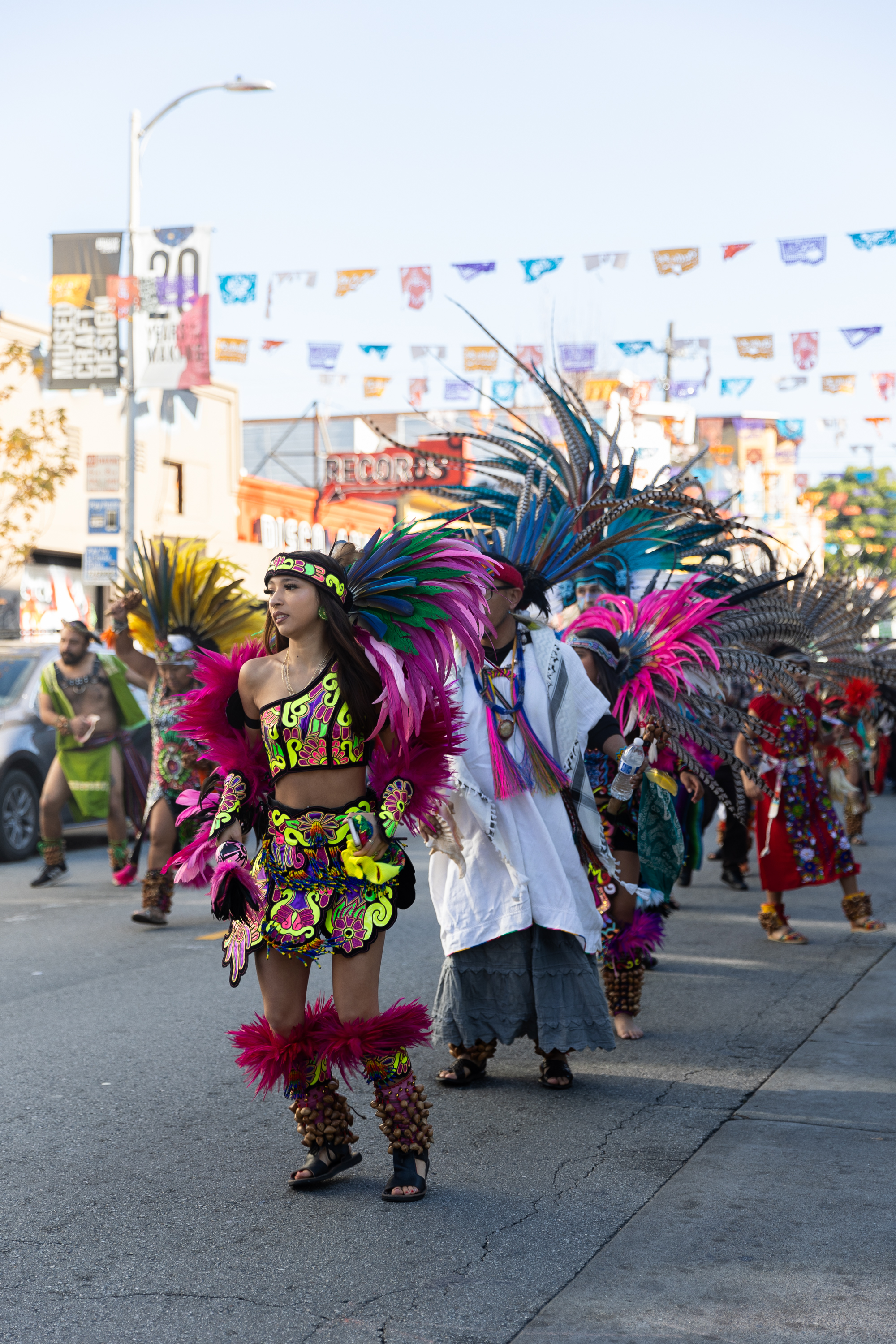 People parade in vibrant traditional costumes with colorful feather headdresses on a street, under festive banners strung across.