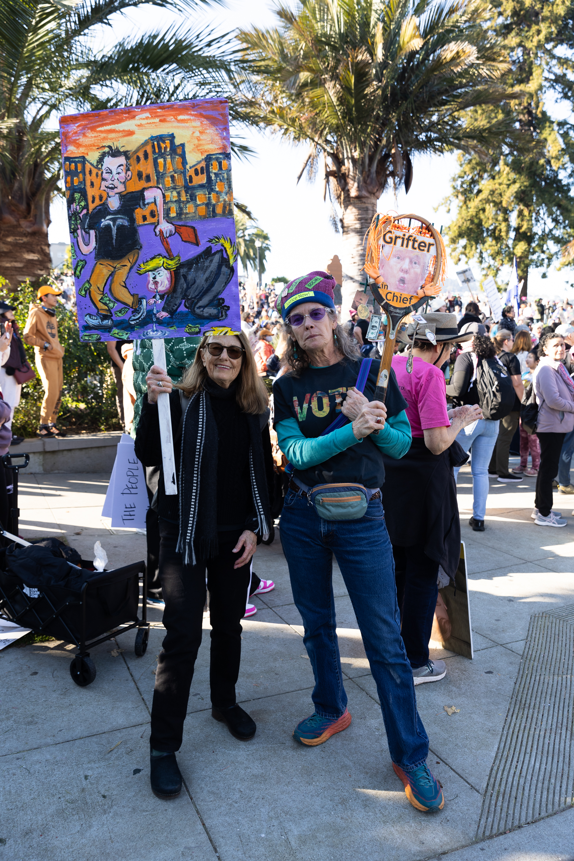 Two women hold protest signs amid a crowd, with palm trees in the background. One sign features a colorful illustration, and the other includes text.