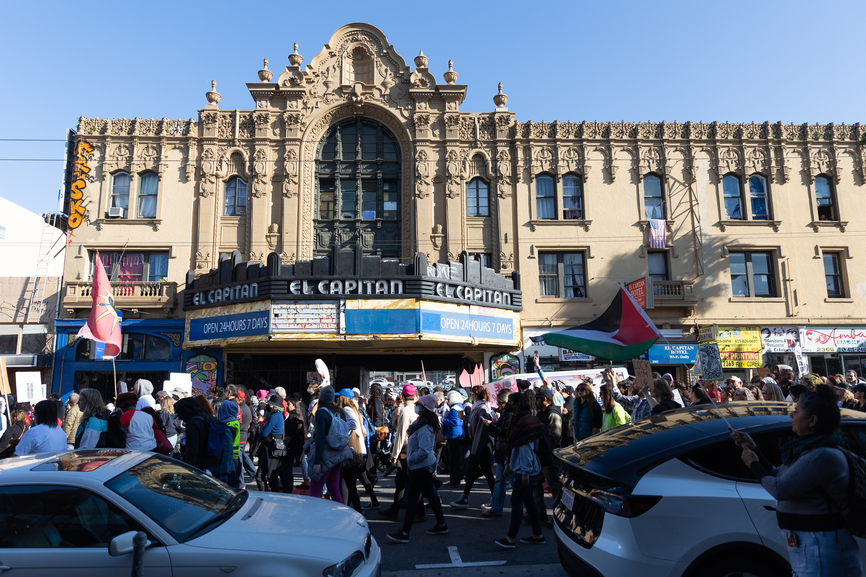 A crowd marches in front of an ornate building labeled &quot;El Capitan.&quot; People hold flags and signs, and there are cars parked on the street.