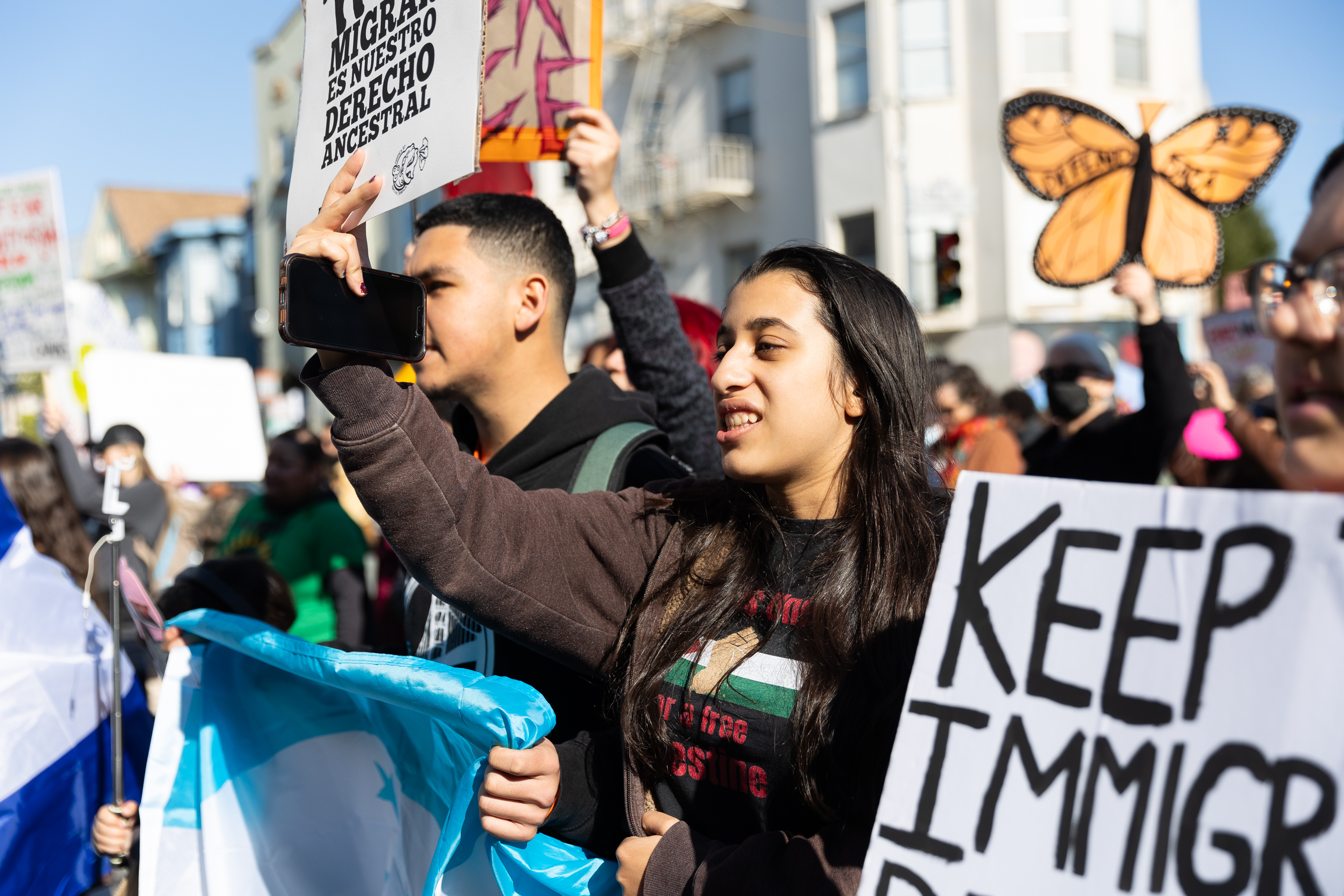 People are gathered in a protest, holding signs with messages about immigration rights and social justice. A butterfly-shaped sign is visible in the background.