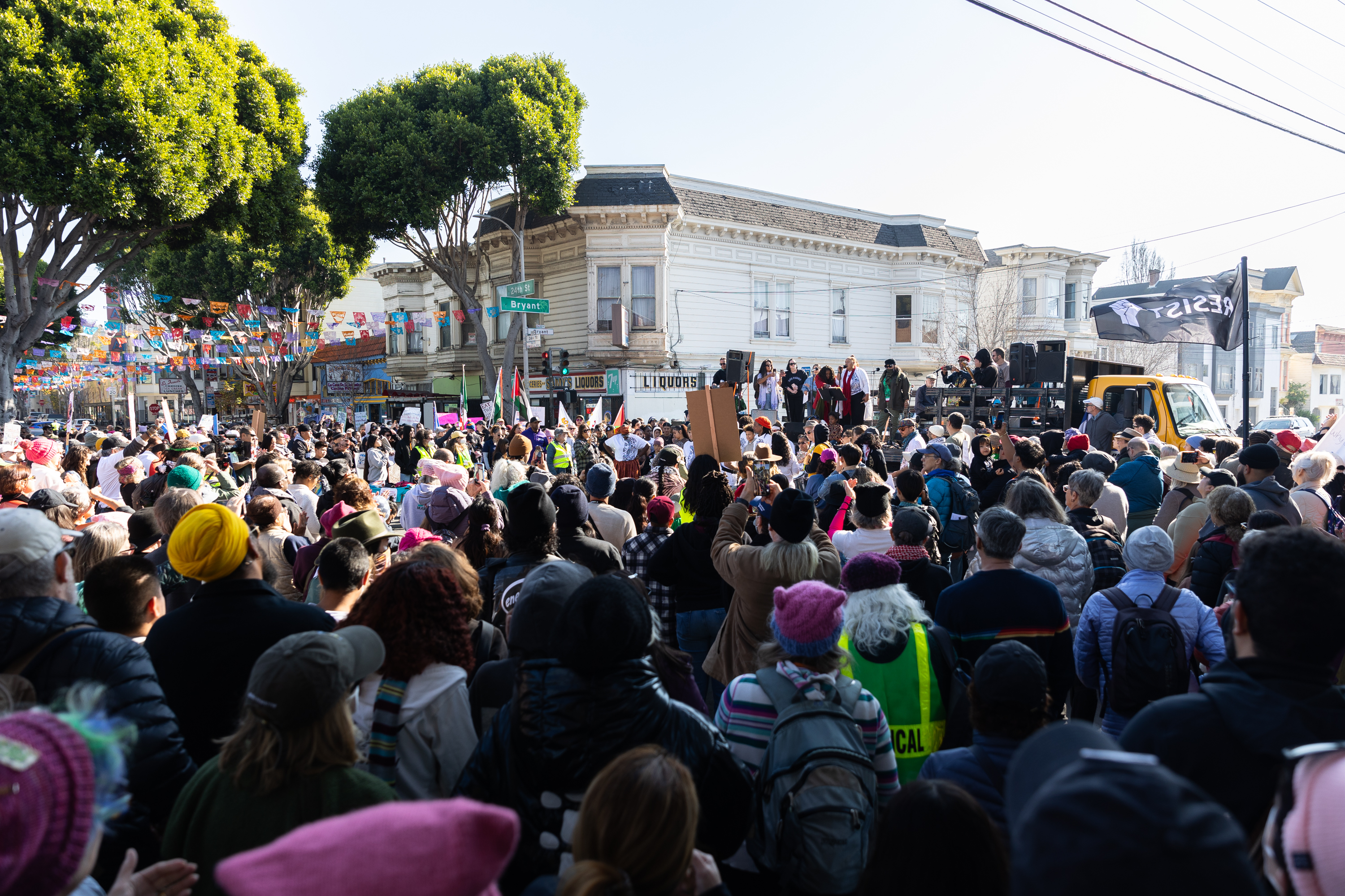 A large crowd gathers in a street for a rally or protest. People hold signs near a stage with speakers. Colorful flags hang above, and trees and buildings surround the scene.