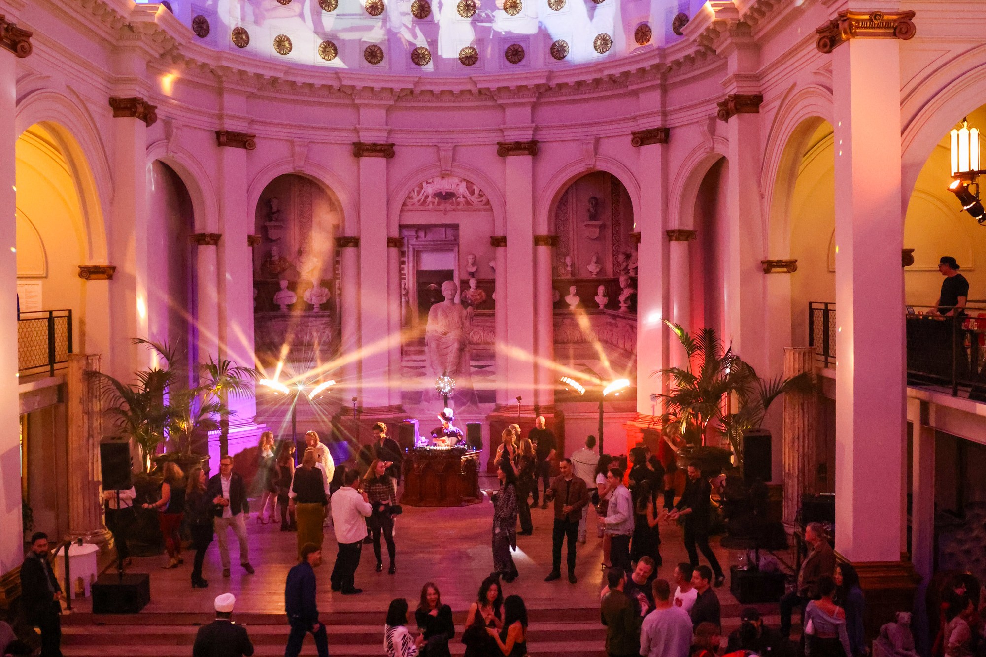 A vibrant party in a grand hall features a domed ceiling, colorful lights, and people mingling and dancing. A DJ is set up on a stage surrounded by ornate arches.