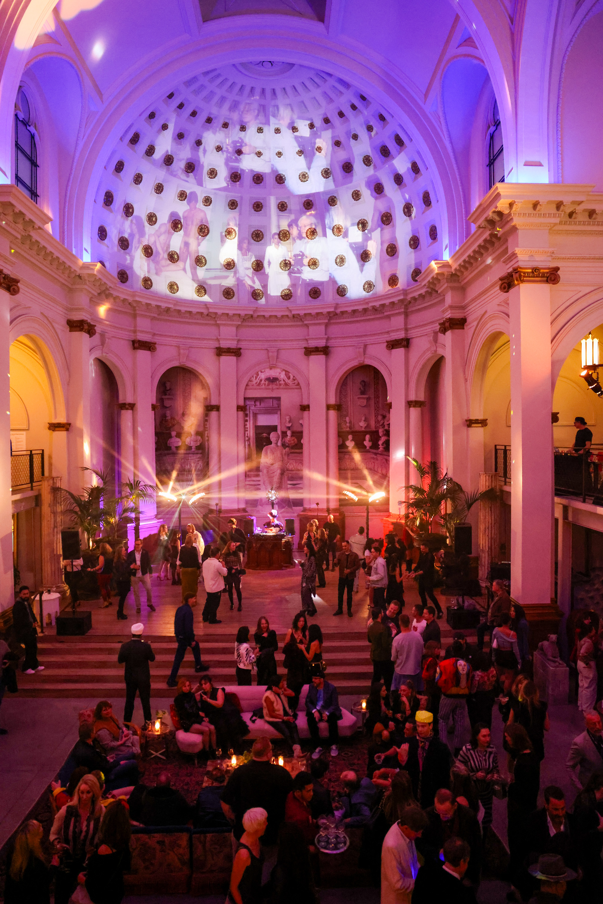 A vibrant party in a grand hall features a domed ceiling, colorful lights, and people mingling and dancing. A DJ is set up on a stage surrounded by ornate arches.