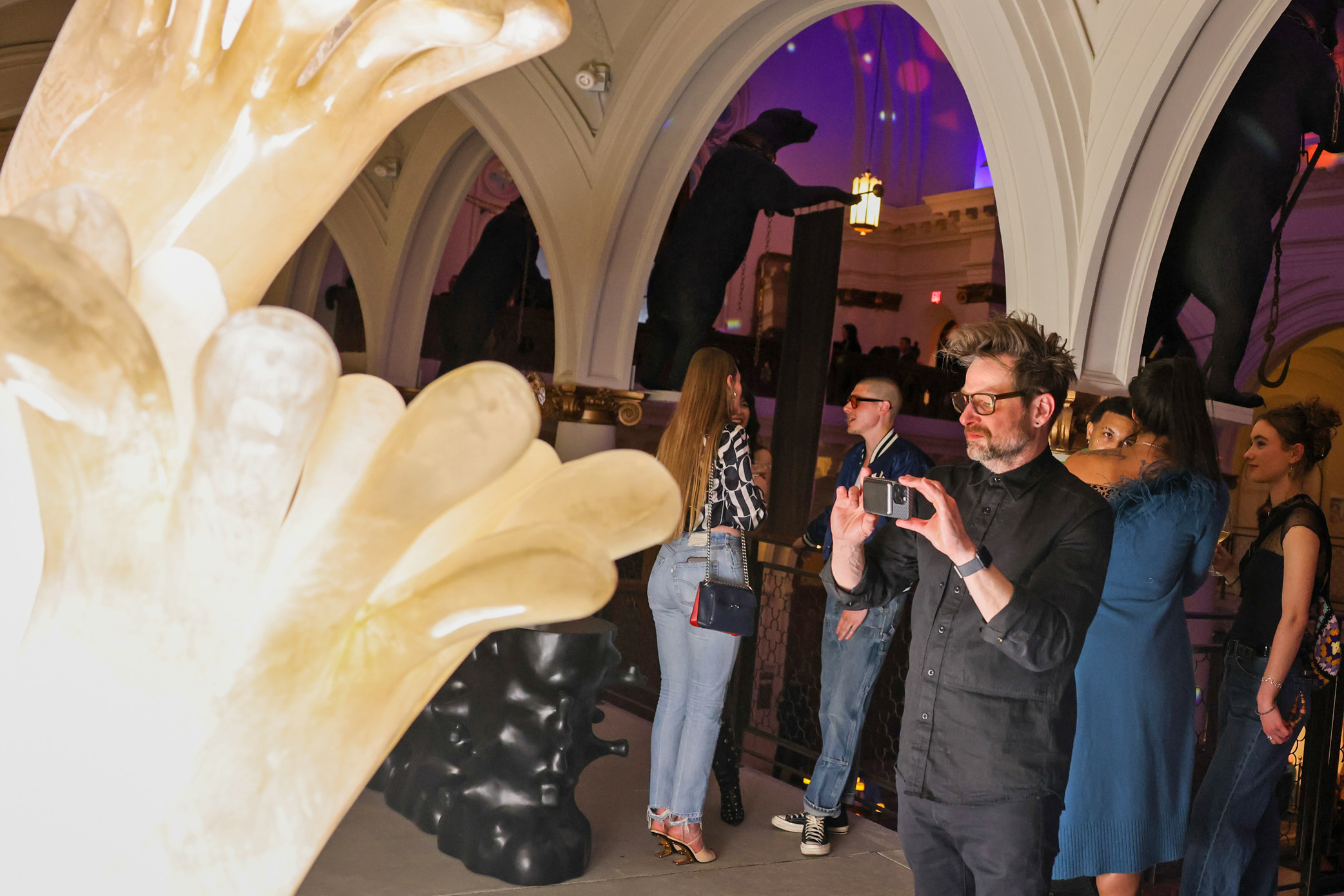 A man takes a photo with his phone in an art gallery, featuring an abstract sculpture in the foreground. Several people stand and chat under arched ceilings.