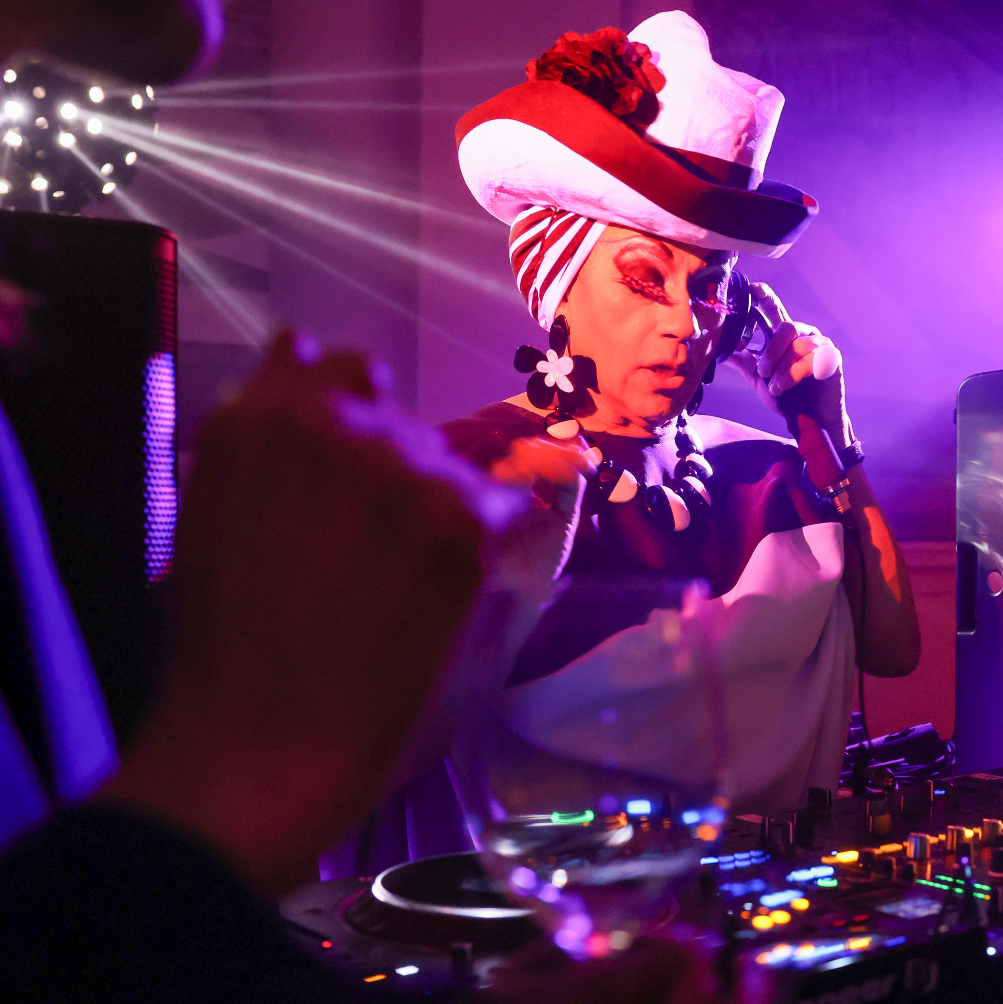 A DJ in a vibrant setting wears a striking red and white hat, bold makeup, and large earrings. Colorful stage lights illuminate the scene, creating a lively atmosphere.