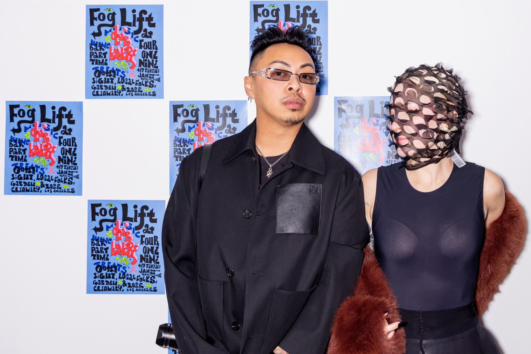 Two people stand in front of a wall with posters saying &quot;Fog Lift.&quot; One person wears a black outfit and glasses, the other has a netted face cover and fur shawl.