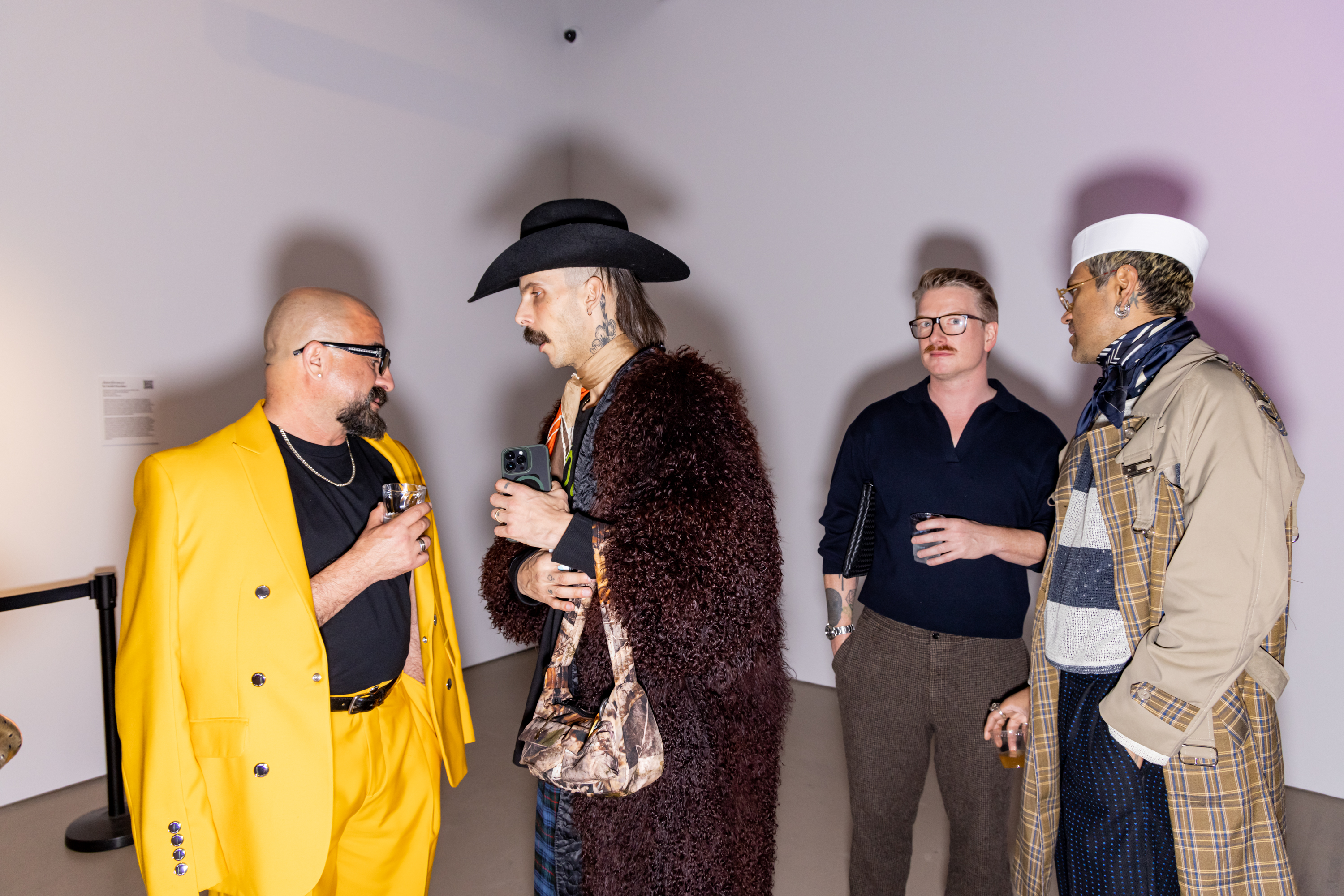 Four people in conversation at an event. One wears a bright yellow suit, another a furry coat and cowboy hat, the others casual; drinks in hand, text on wall.