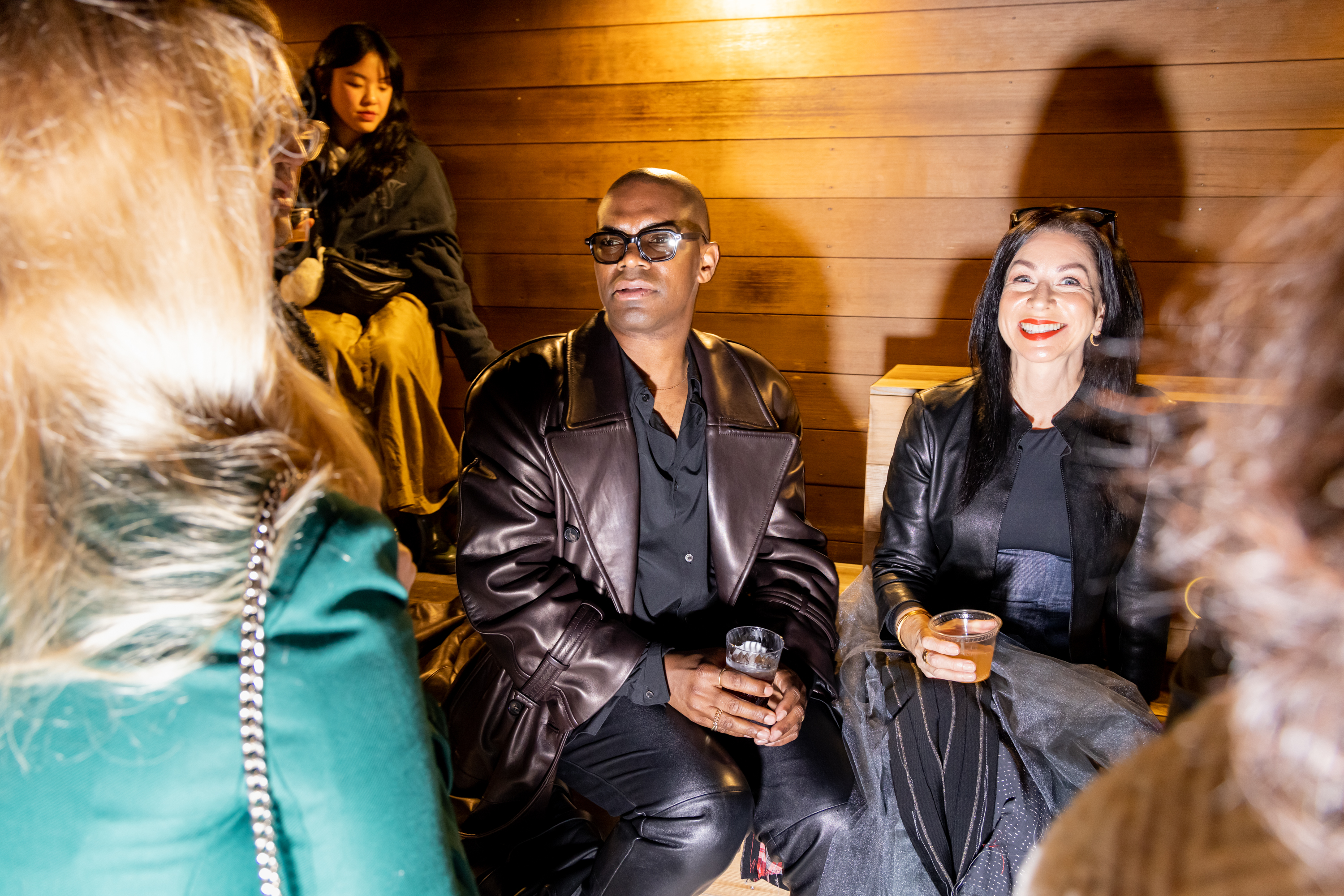 A group is gathered indoors. A person in sunglasses and leather sits beside a smiling woman with dark hair, each holding a drink, while others chat nearby.