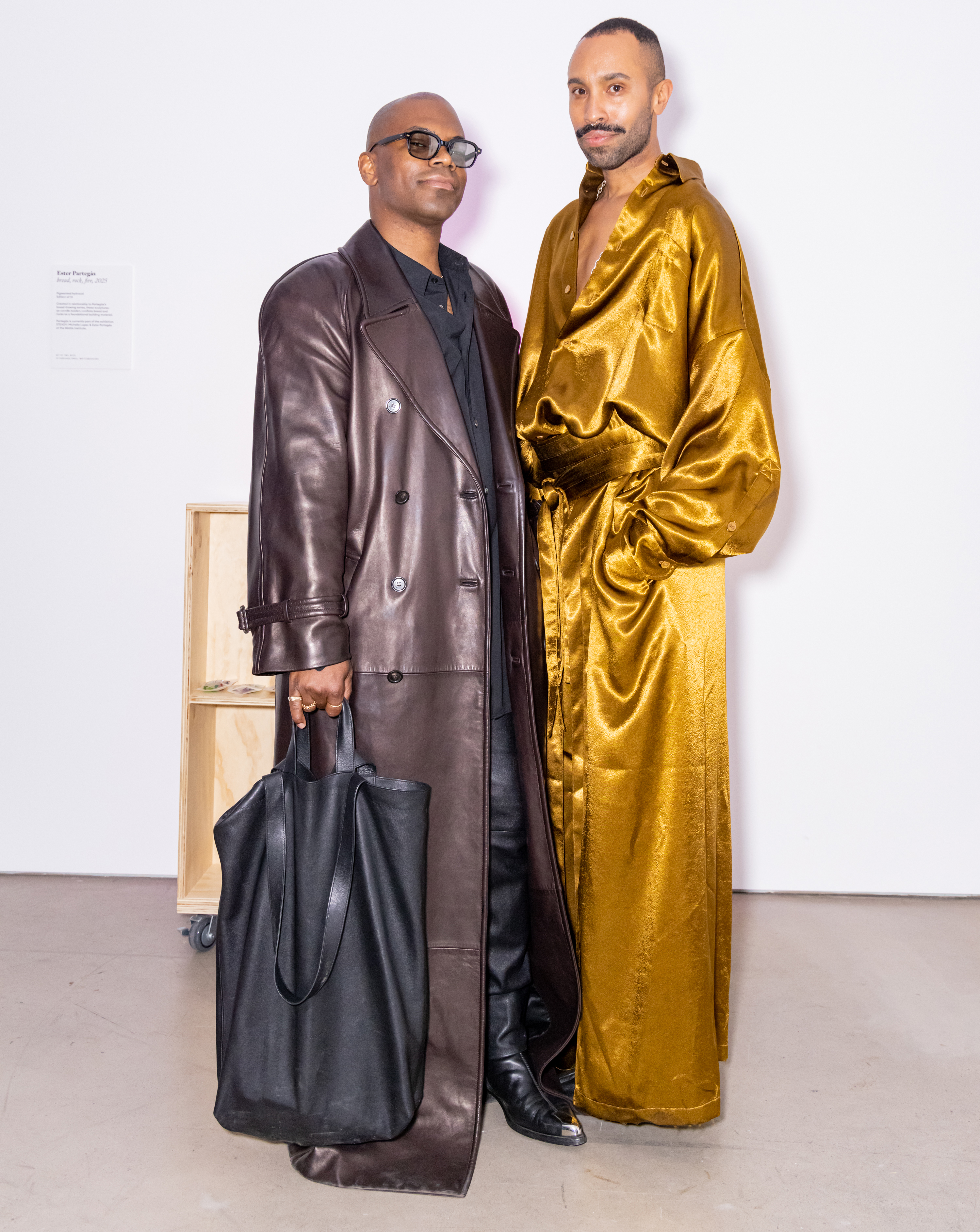 Two individuals pose together; one wears a long brown leather coat and carries a black bag, while the other is in a shiny gold robe.