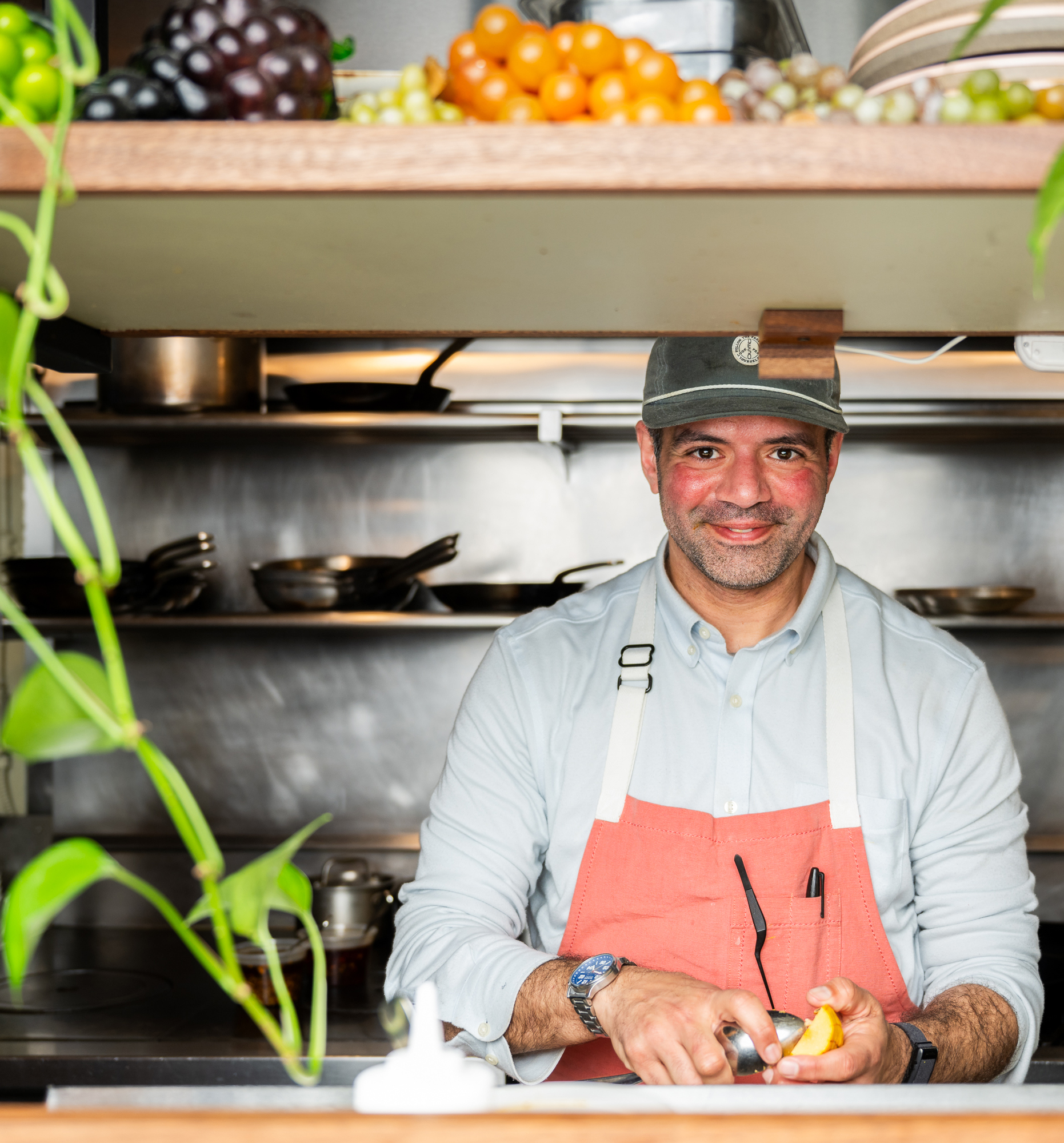 A smiling person in an apron peels an orange at a kitchen counter, with shelves of cookware and fresh fruit above. Green plants add a touch of color.