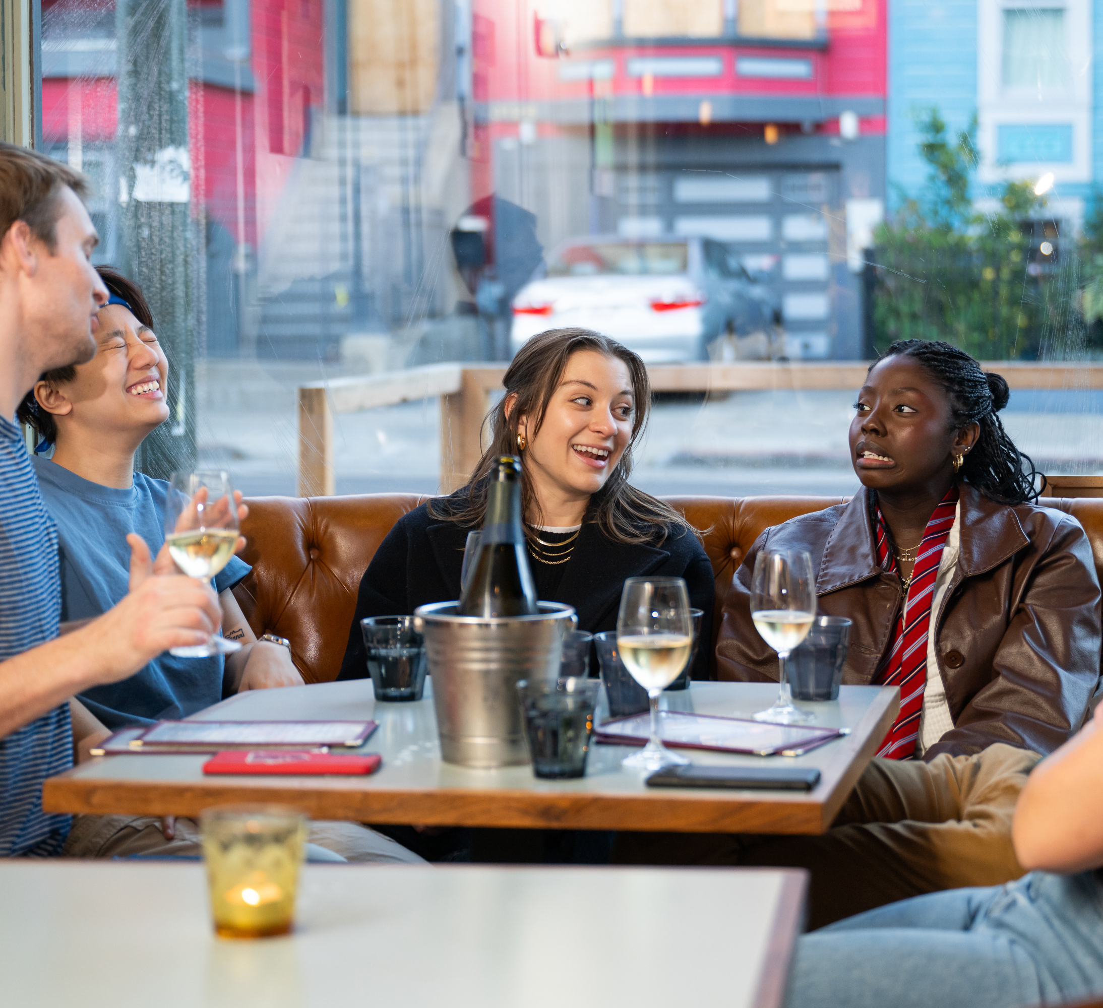 Four people sit around a table in a restaurant, laughing and talking. There are drinks in front of them and a bucket with a bottle, with a street view outside.