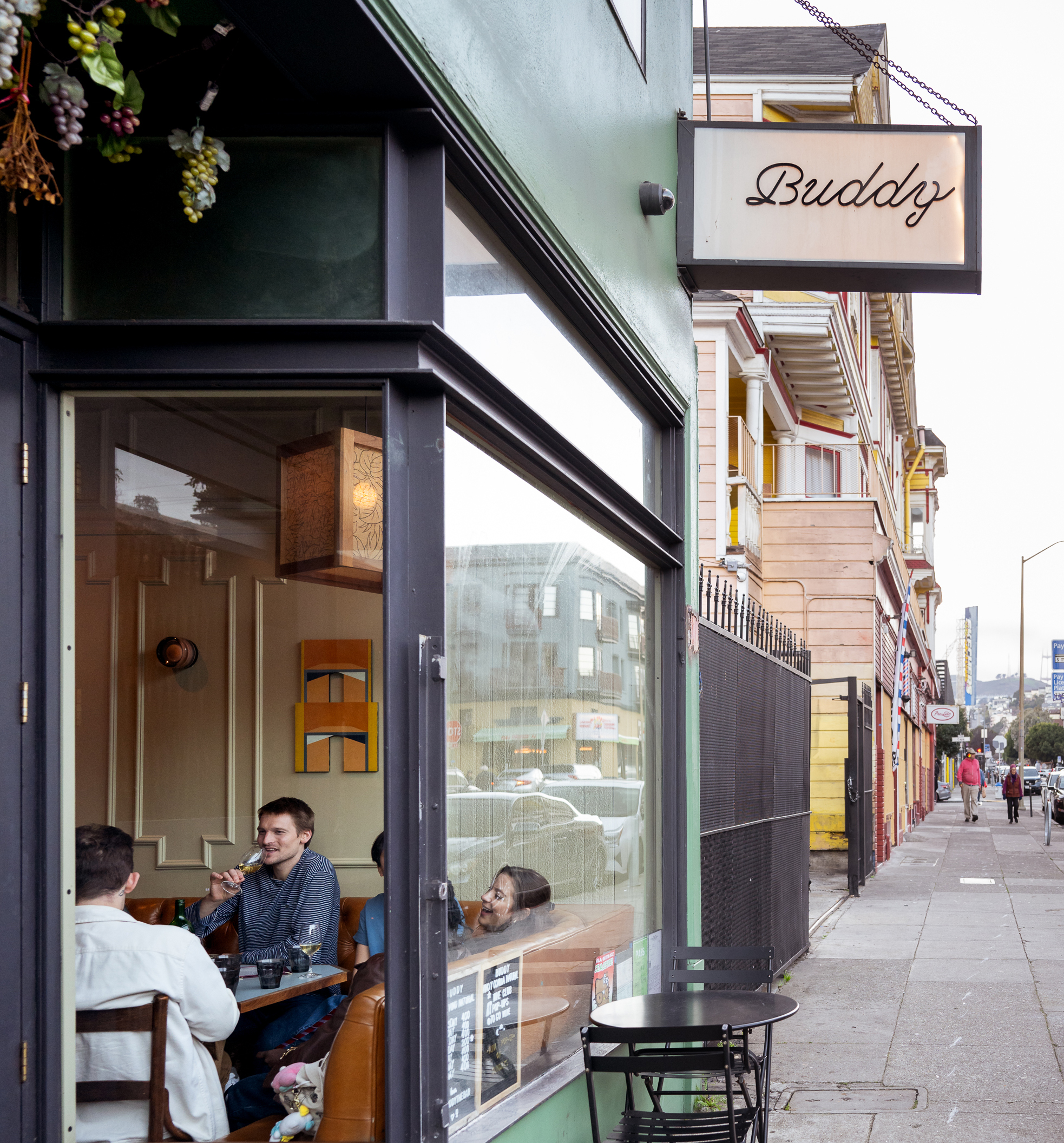 A cozy cafe named Buddy on a city street. Inside the window, three people are seated at a table, enjoying a conversation. Outside, a small round table sits on the sidewalk.