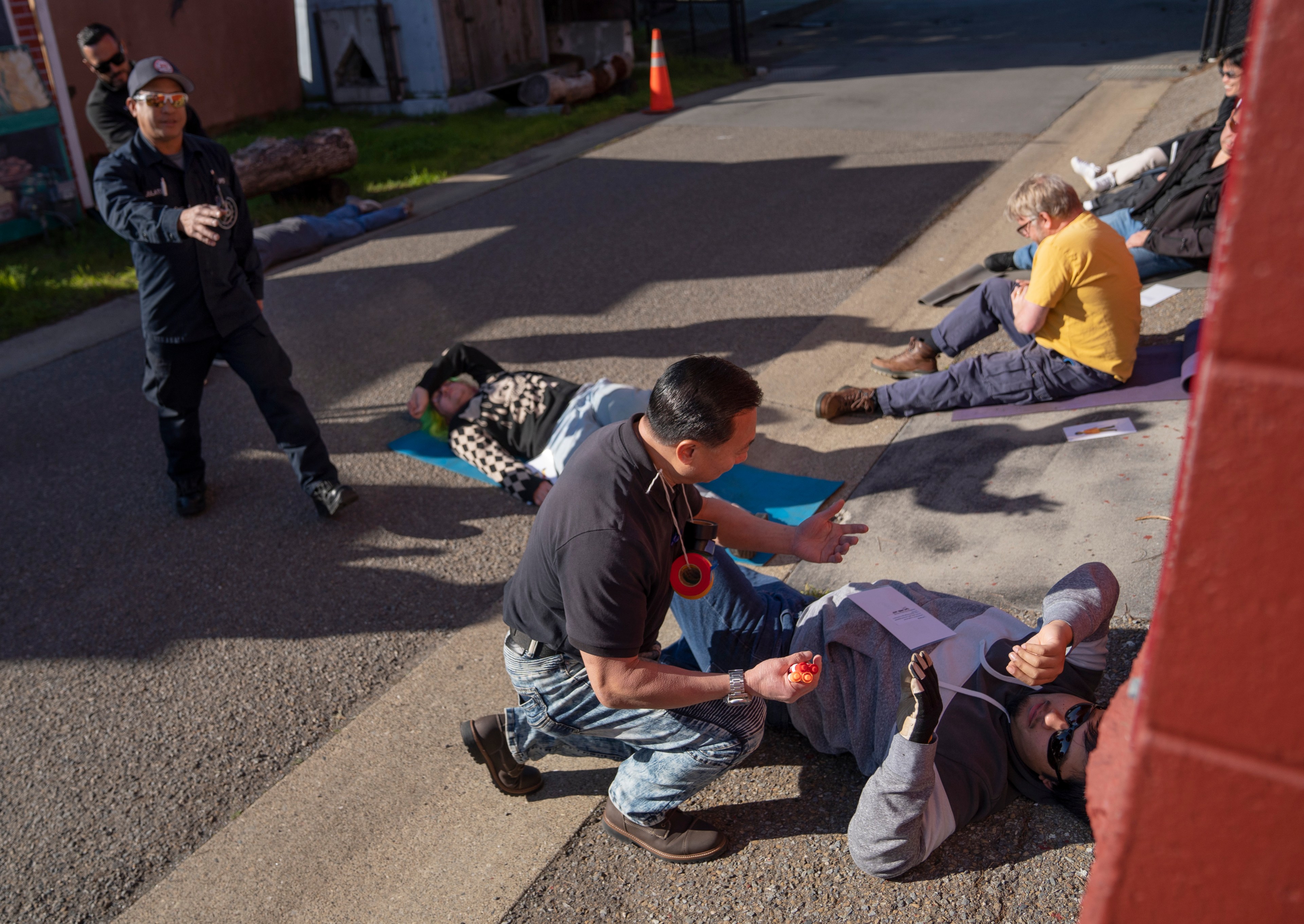 People are involved in a first aid training scenario on a sunny street. Some participants are lying down, while others kneel beside them, offering assistance.