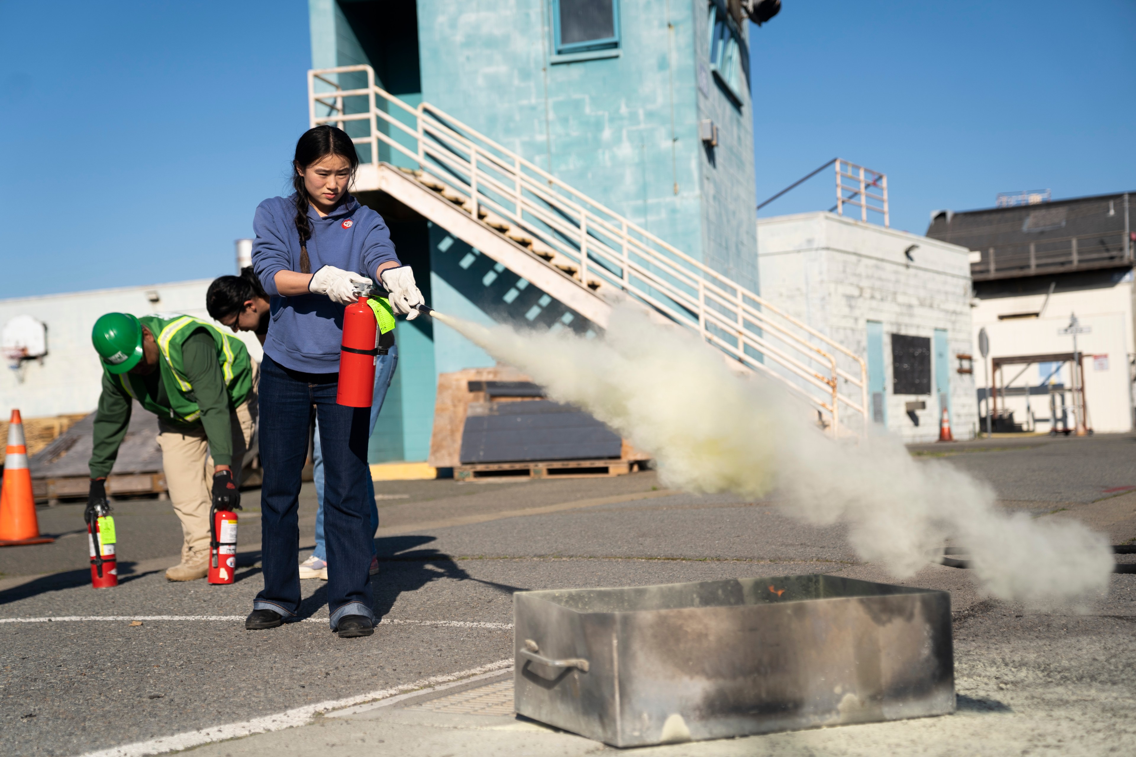 A woman in gloves uses a fire extinguisher, spraying foam towards a metal tray on the ground. Two other people with extinguishers are nearby outside a blue building.