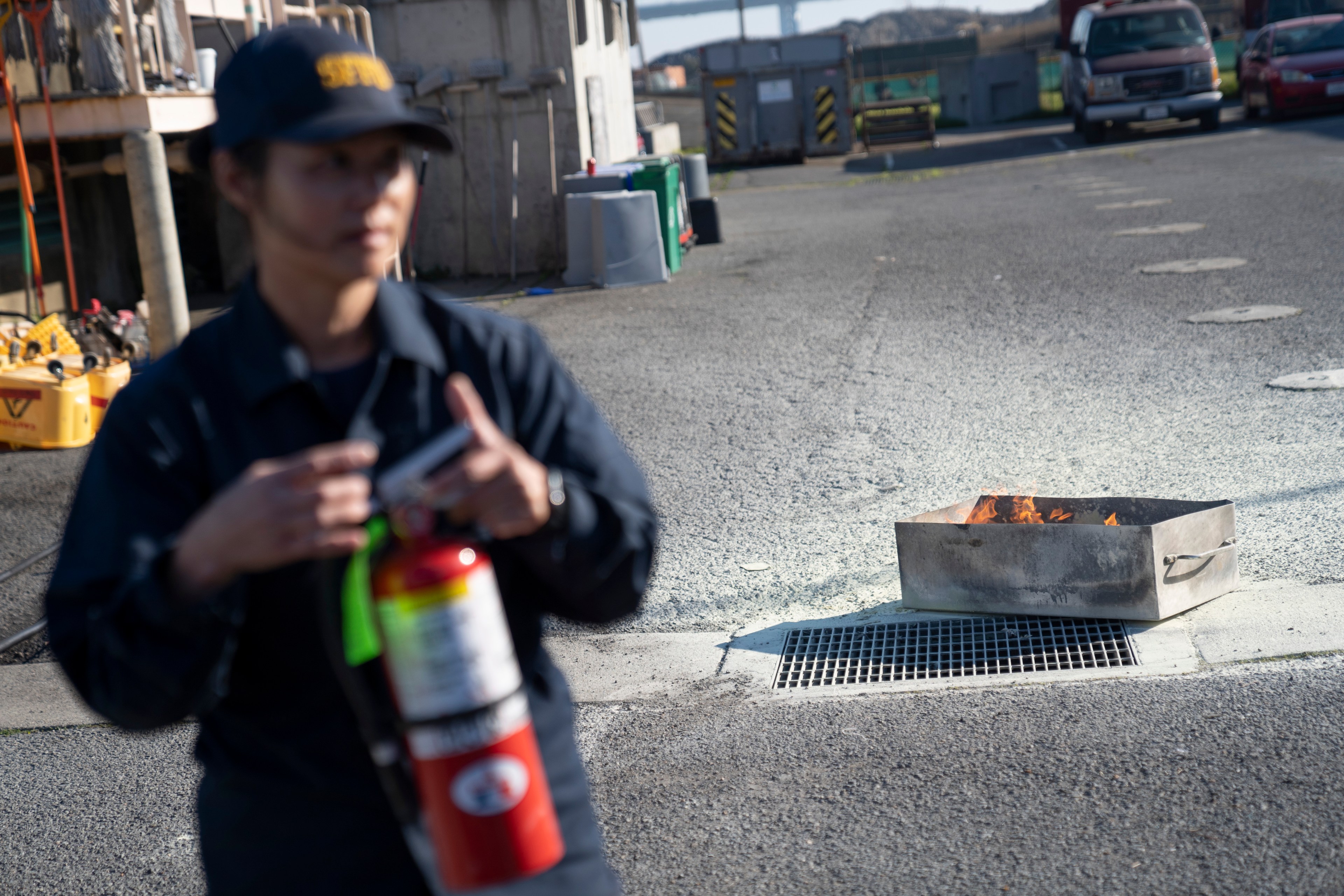 A person holding a fire extinguisher is in the foreground, slightly blurred. In the background, there is a metal container on the ground with flames inside.