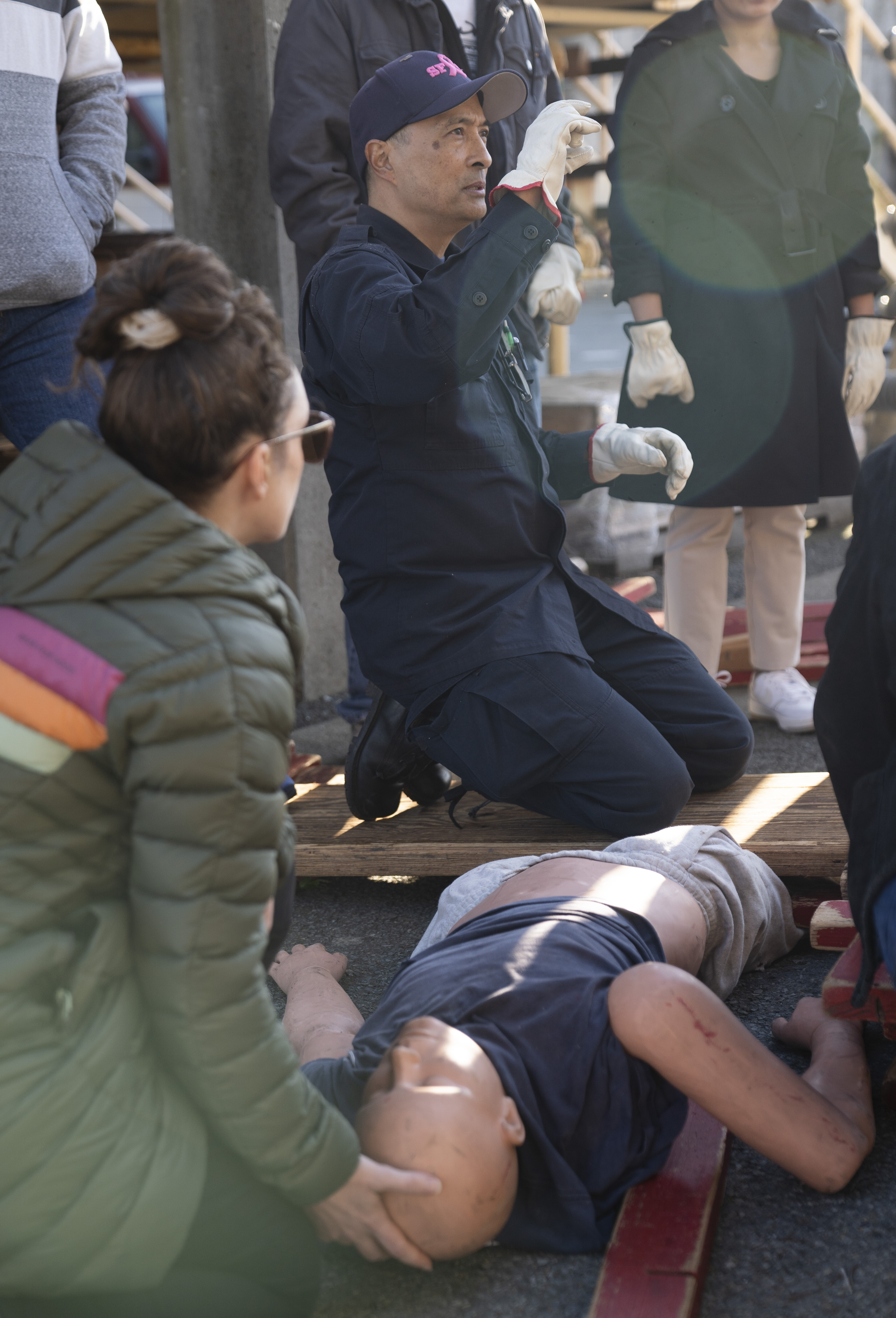 A man wearing gloves and a cap kneels beside a mannequin on the ground. He appears to be instructing a group, with a woman holding the mannequin's head.