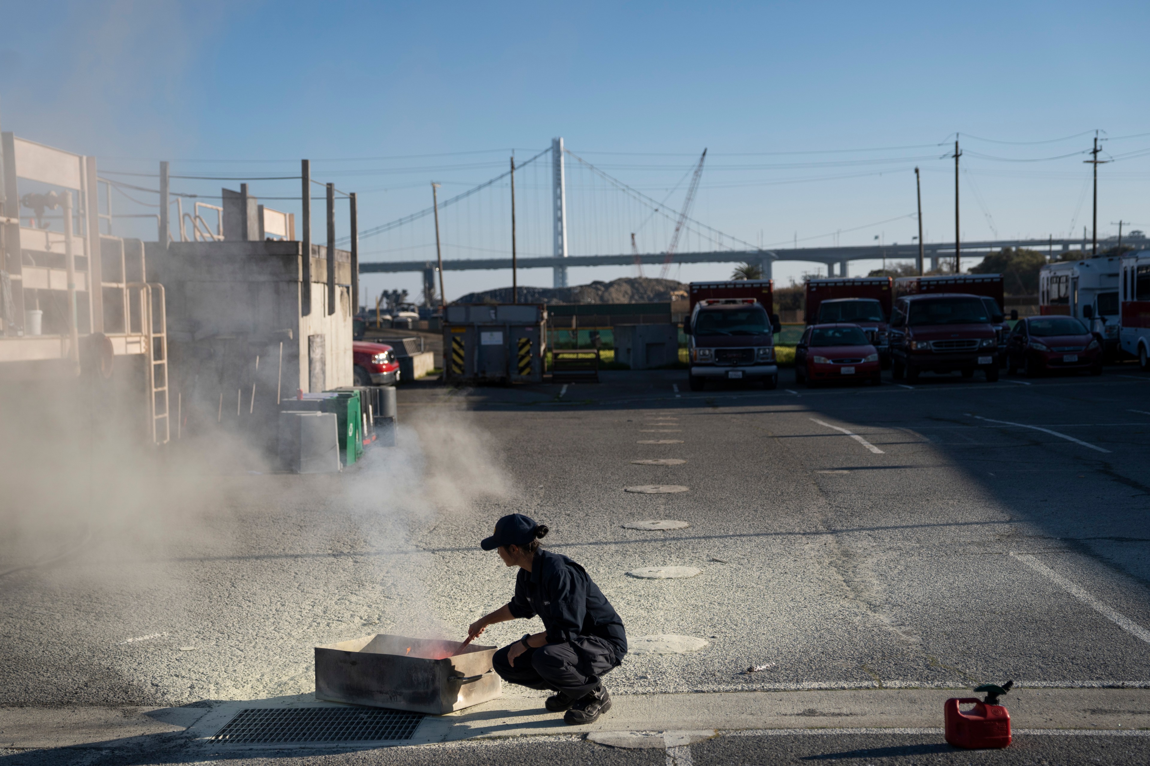 A person in a dark uniform crouches by a smoking fire in a metal tray on a concrete surface, near parked vehicles and a bridge in the background.