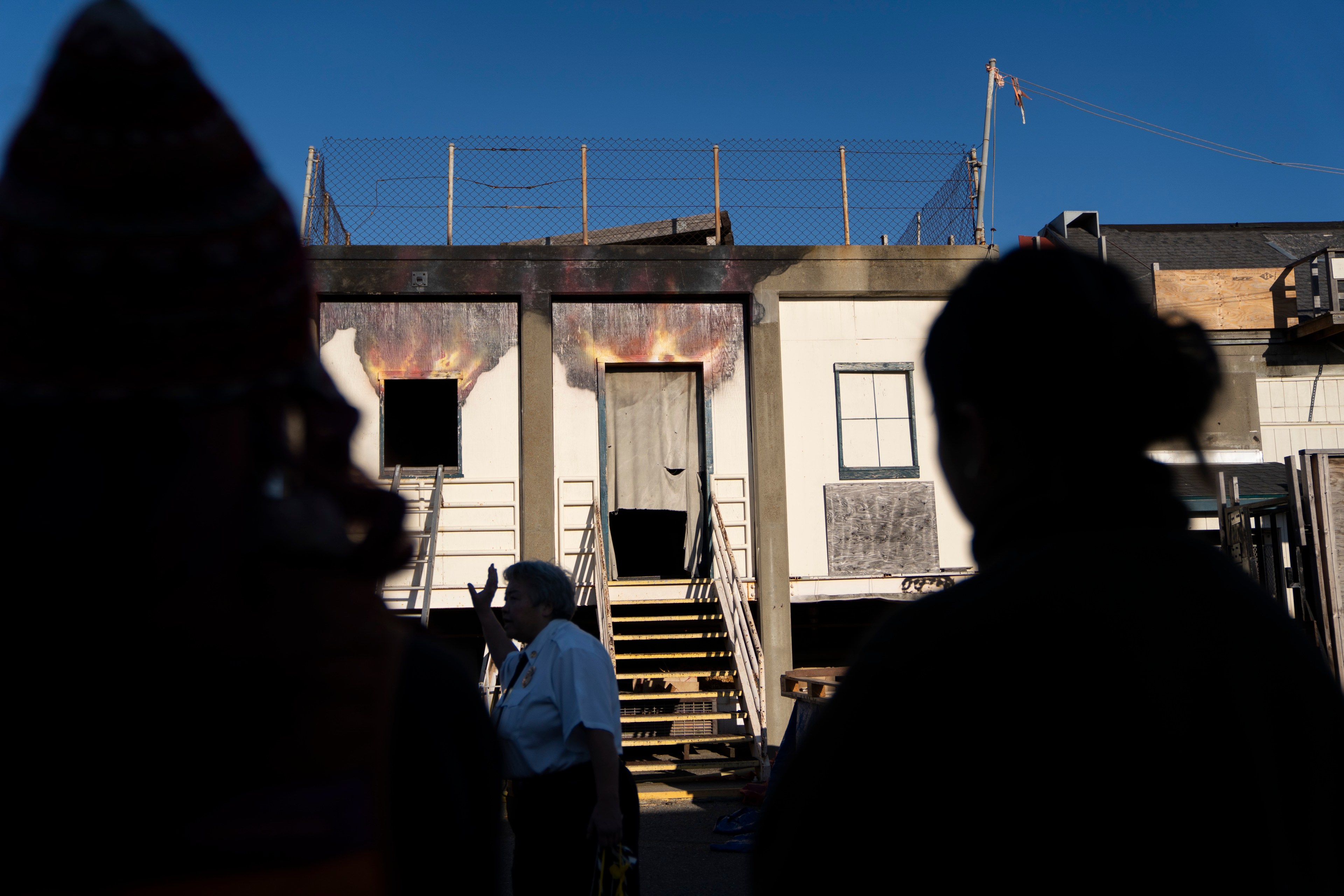 The image shows a silhouette of people observing a burnt-out building with stairs leading to a charred doorway and windows, under a clear blue sky.