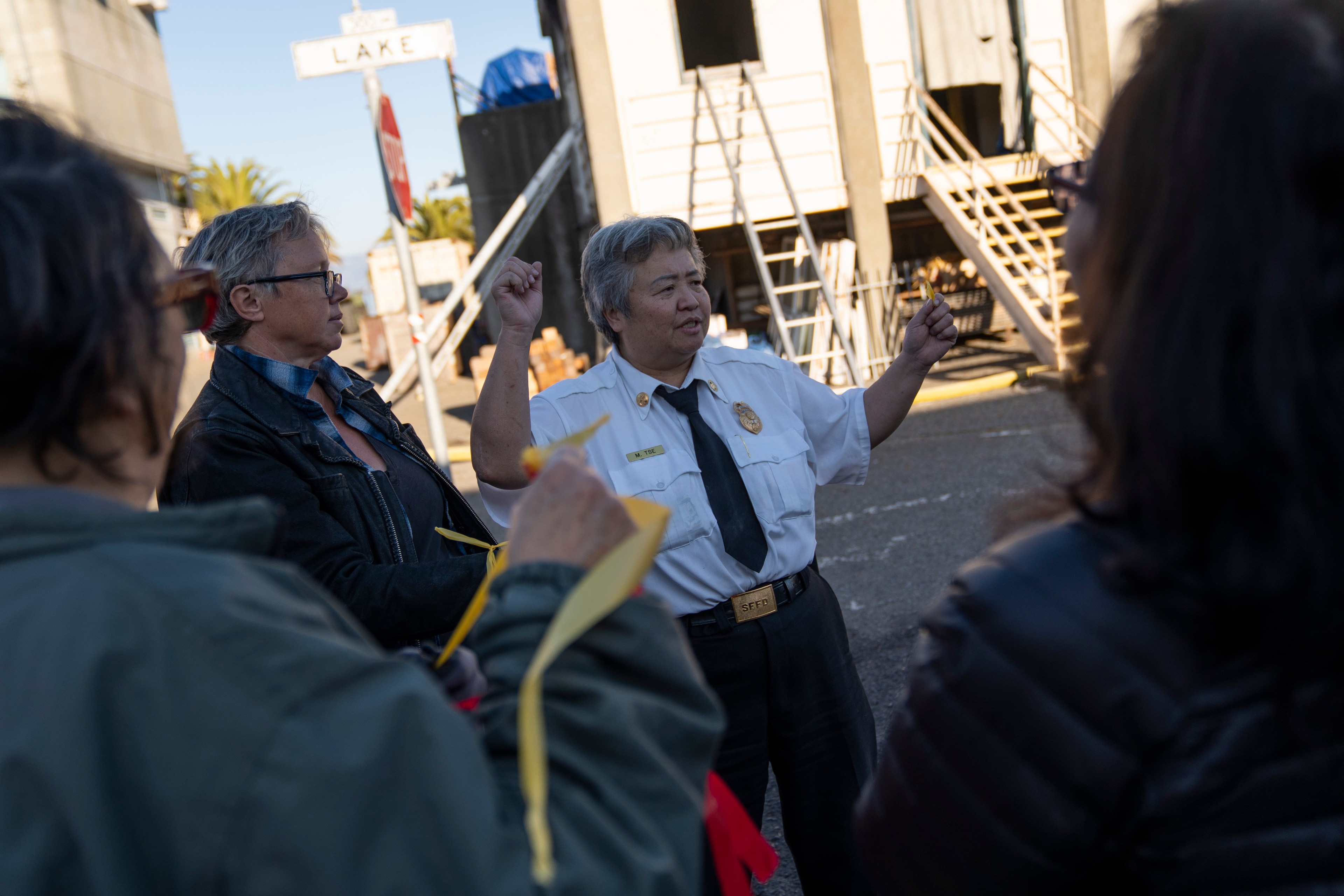 A person in a fire department uniform speaks to a group on a city street. They gesture with both hands, standing in front of a building with staircases.