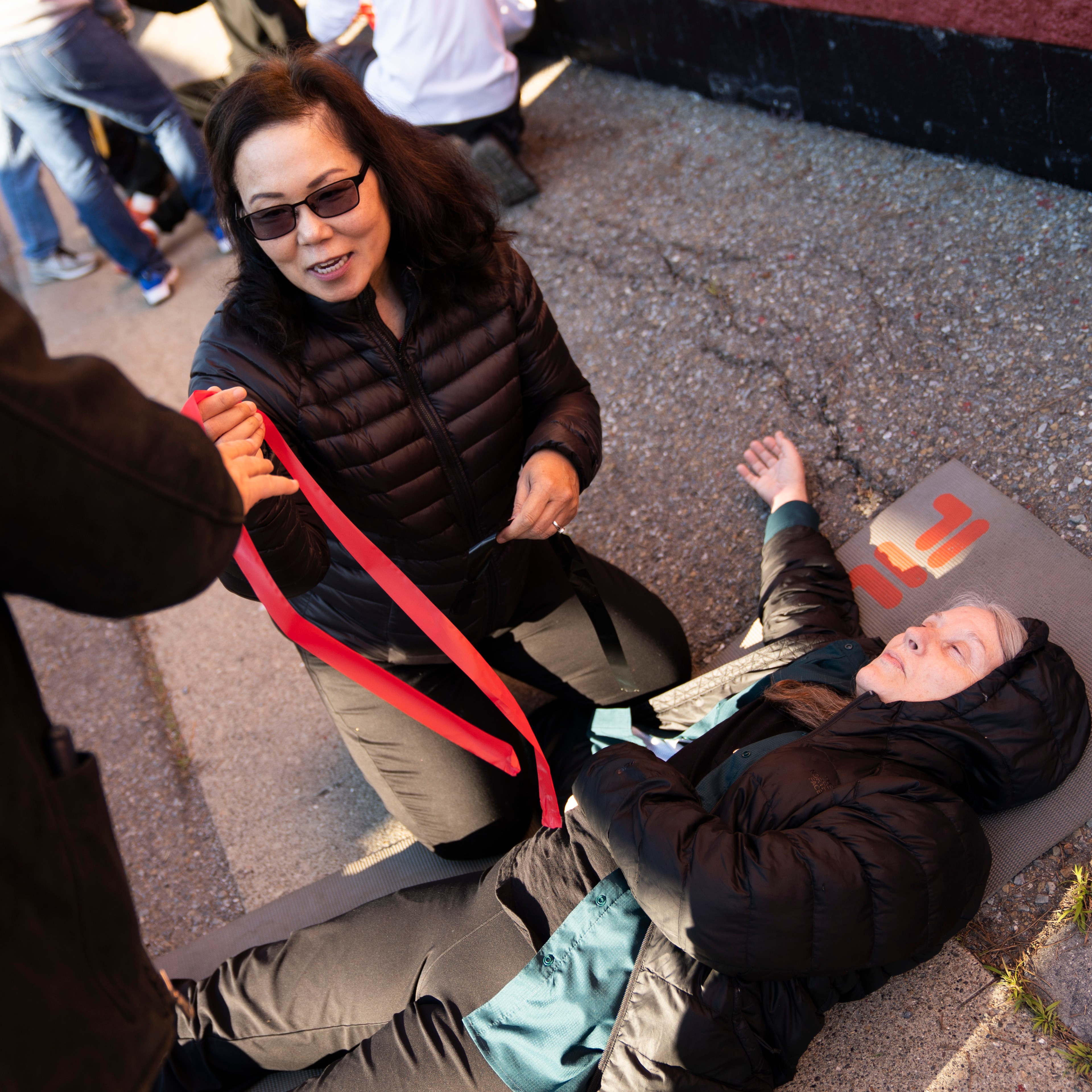 A person in a black jacket kneels, holding a red stripe, next to another person lying on a mat on a concrete surface, wearing a hooded jacket.
