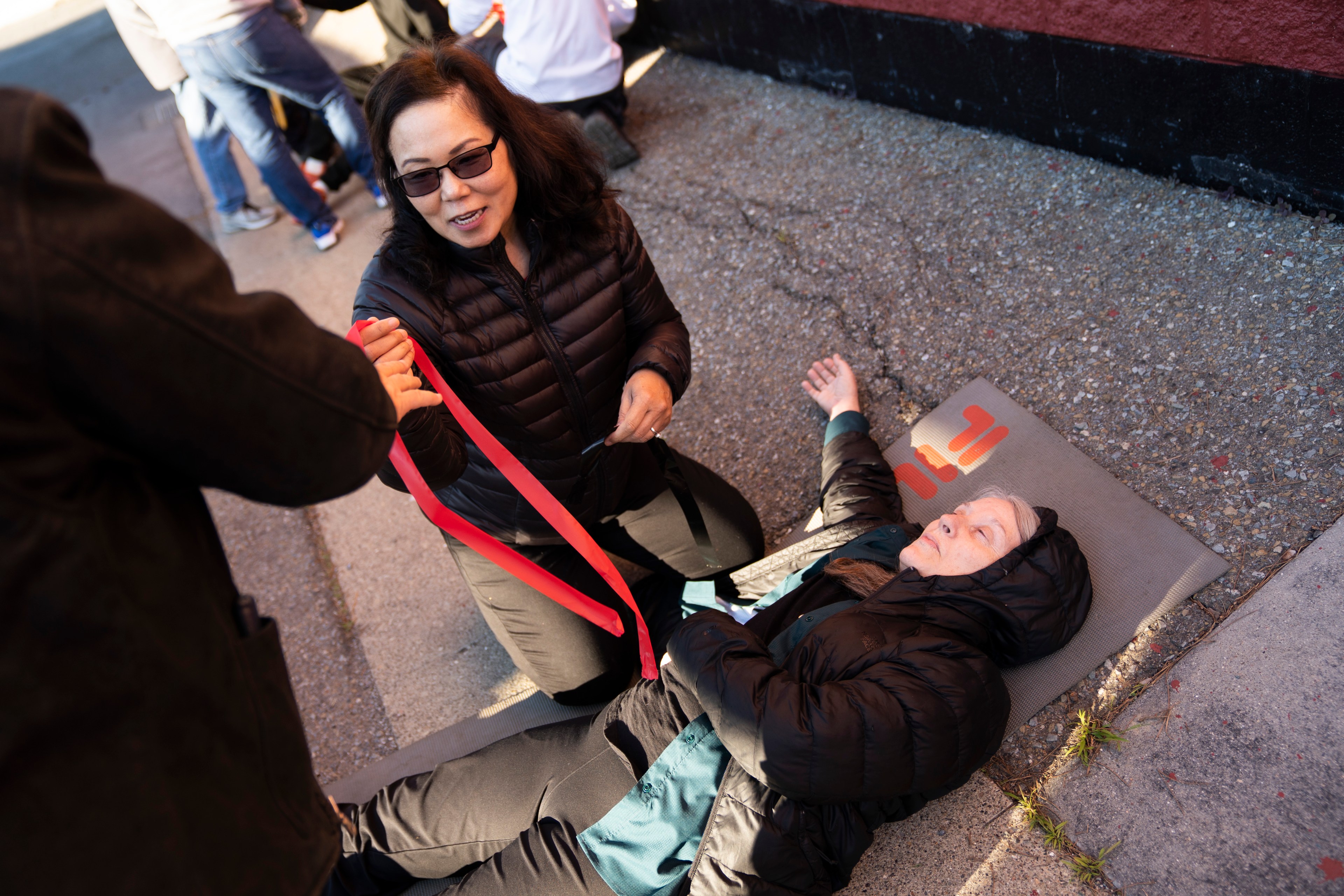 A person in a black jacket kneels, holding a red stripe, next to another person lying on a mat on a concrete surface, wearing a hooded jacket.