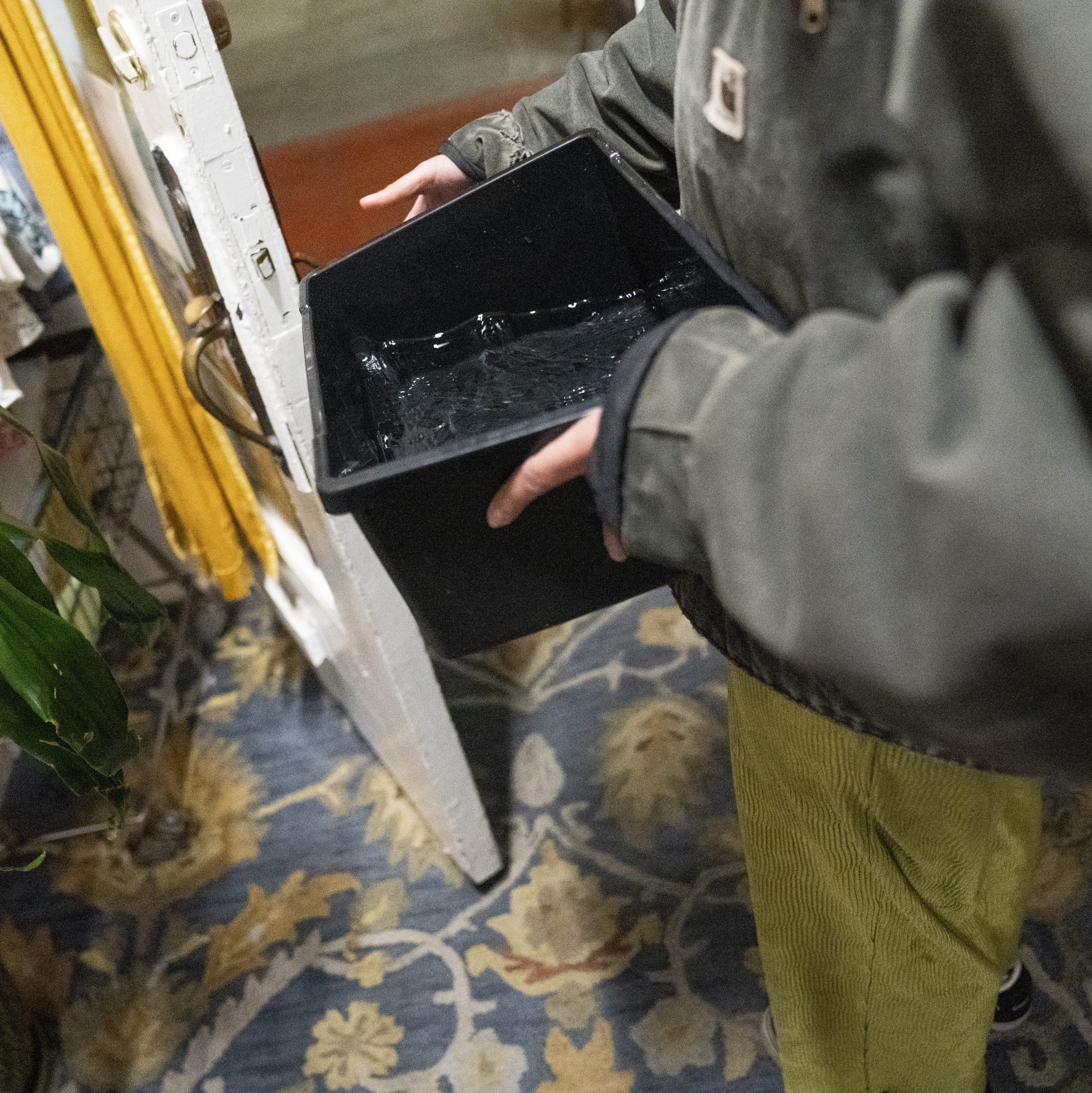 A person in a green jacket holds a black container with water, standing on a floral-patterned rug near a partially opened white door.