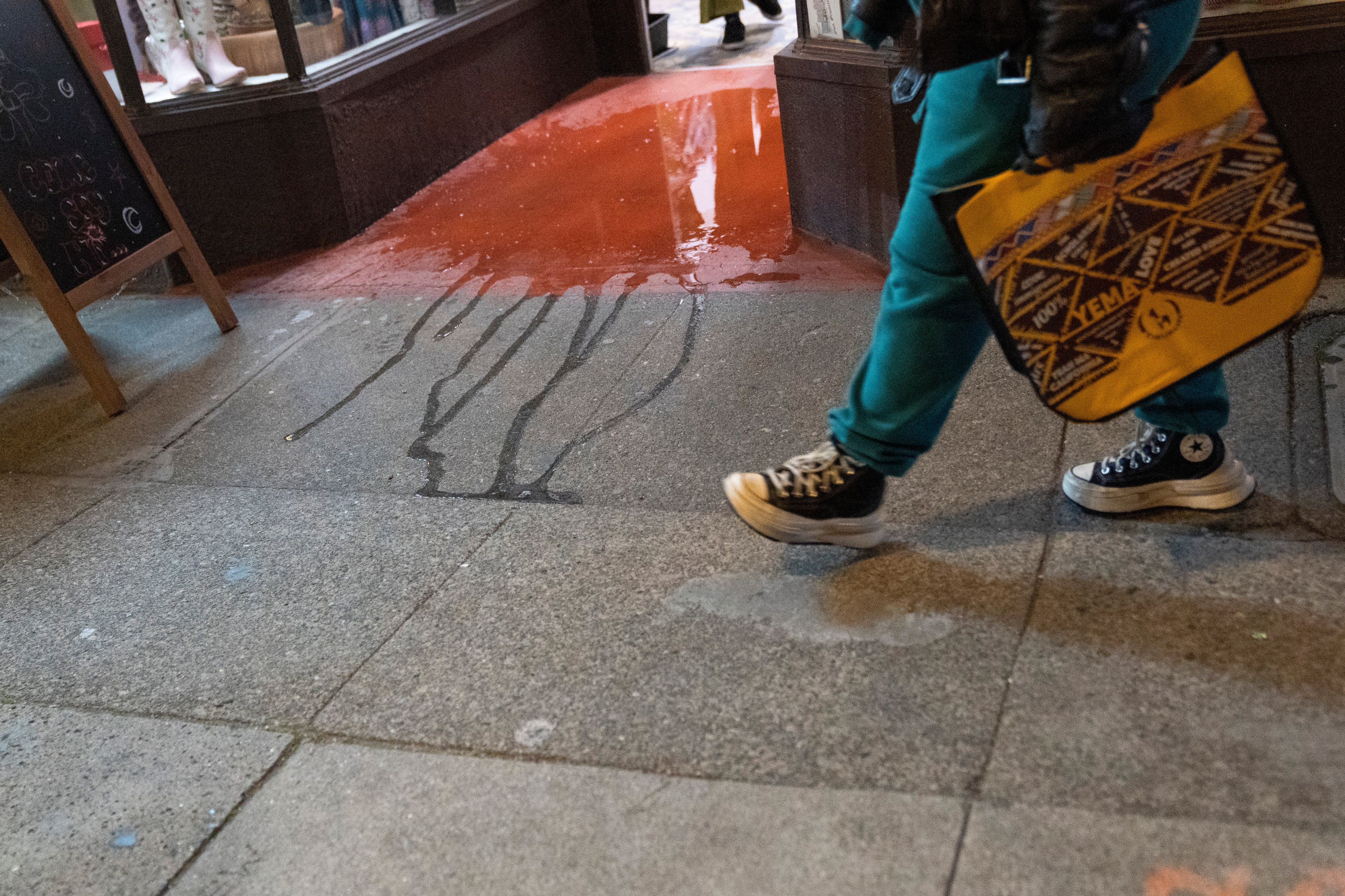 A person walks past a storefront with reddish liquid spilled on the sidewalk. They carry a yellow bag and wear blue pants and black sneakers.