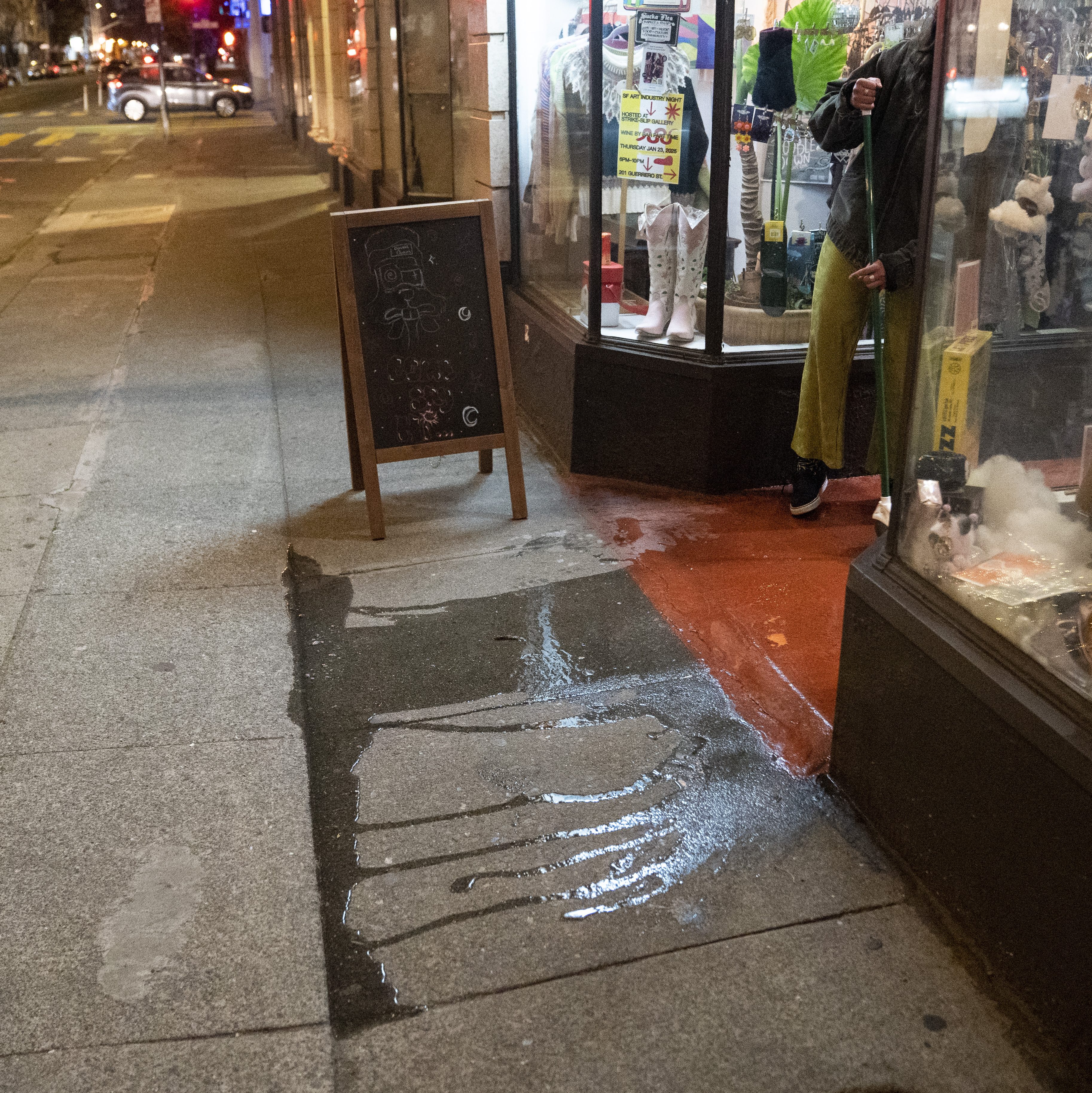 A person in green pants is cleaning a wet sidewalk by a storefront with a chalkboard sign. The area is dimly lit, with various items displayed in the window.