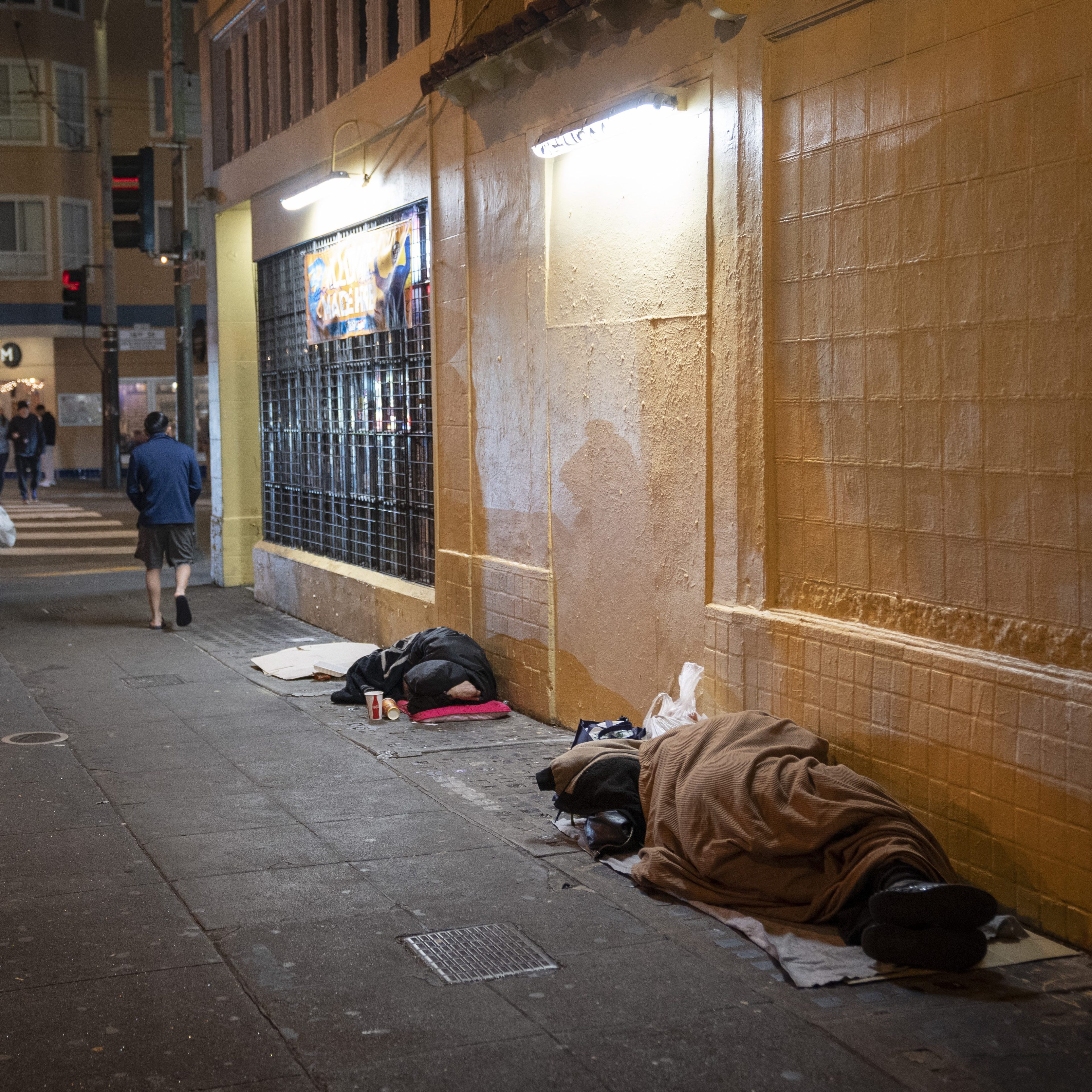 Two people sleep on the sidewalk under blankets beside a building, while a person walks by. A lit sign and a window with bars are visible on the wall.