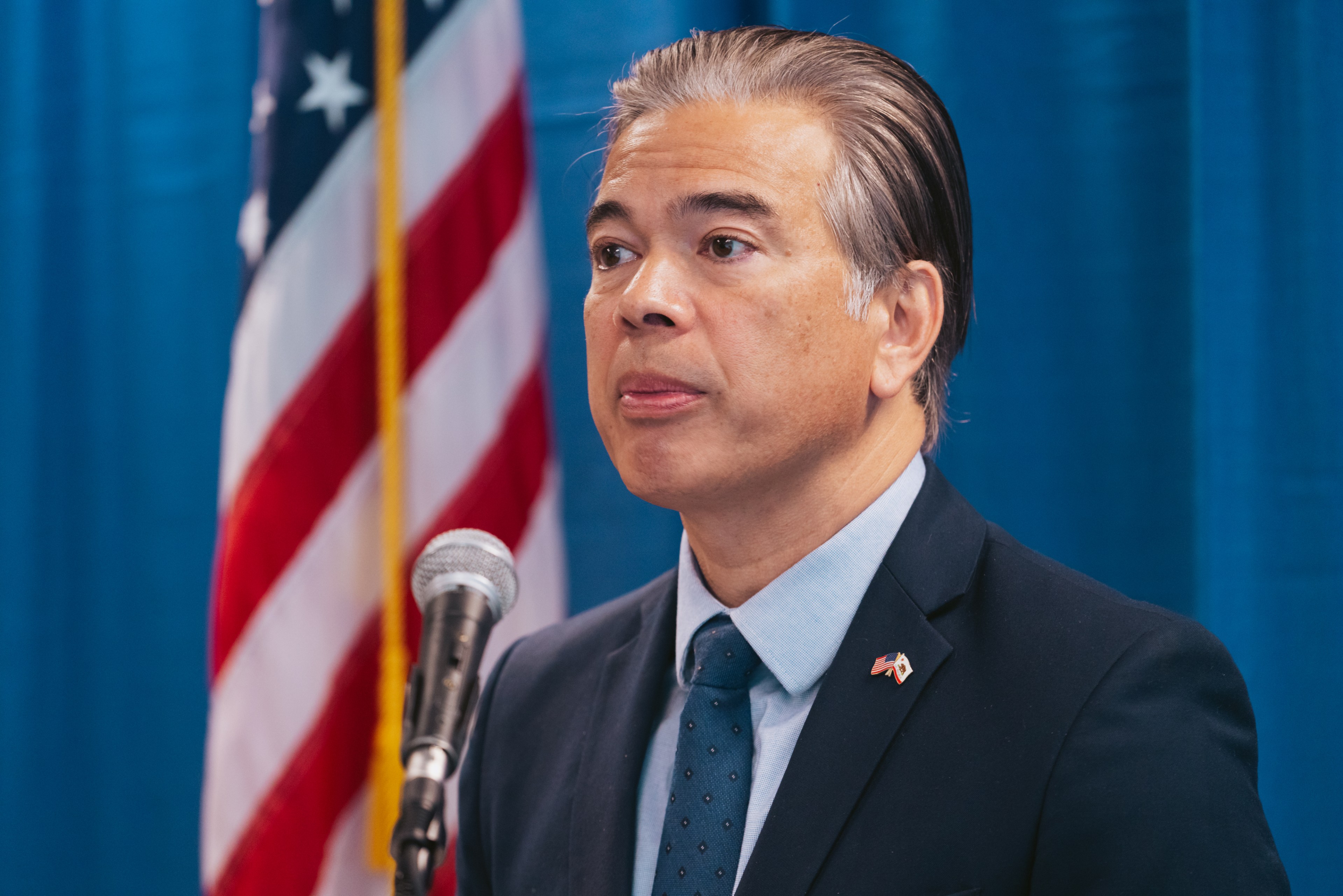 A person in a suit and tie speaks at a microphone. The background is a blue curtain with a U.S. flag visible. He wears a pin with the American flag.