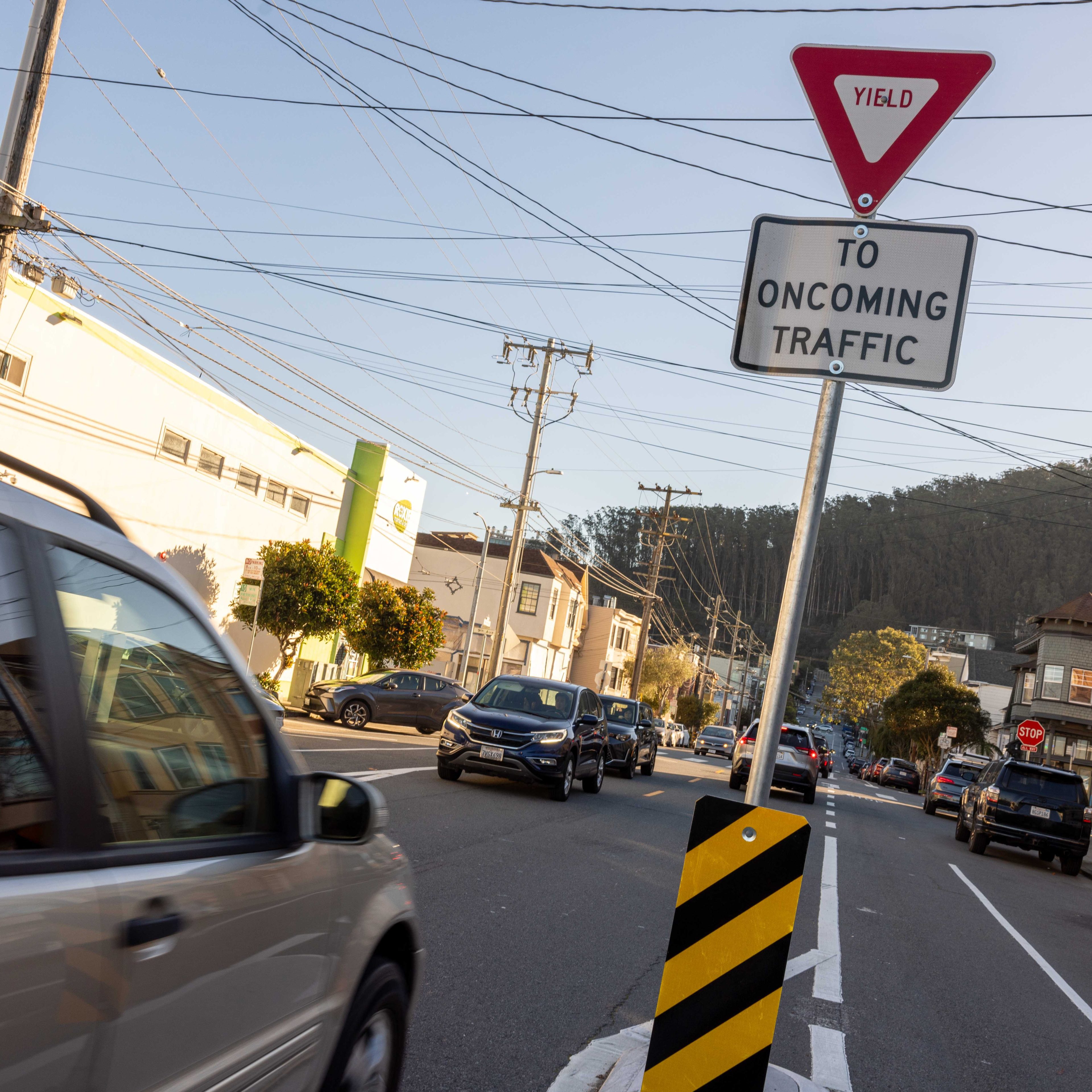 A street with cars passing by has a &quot;Yield to Oncoming Traffic&quot; sign beside a road divider, with buildings and trees in the background.