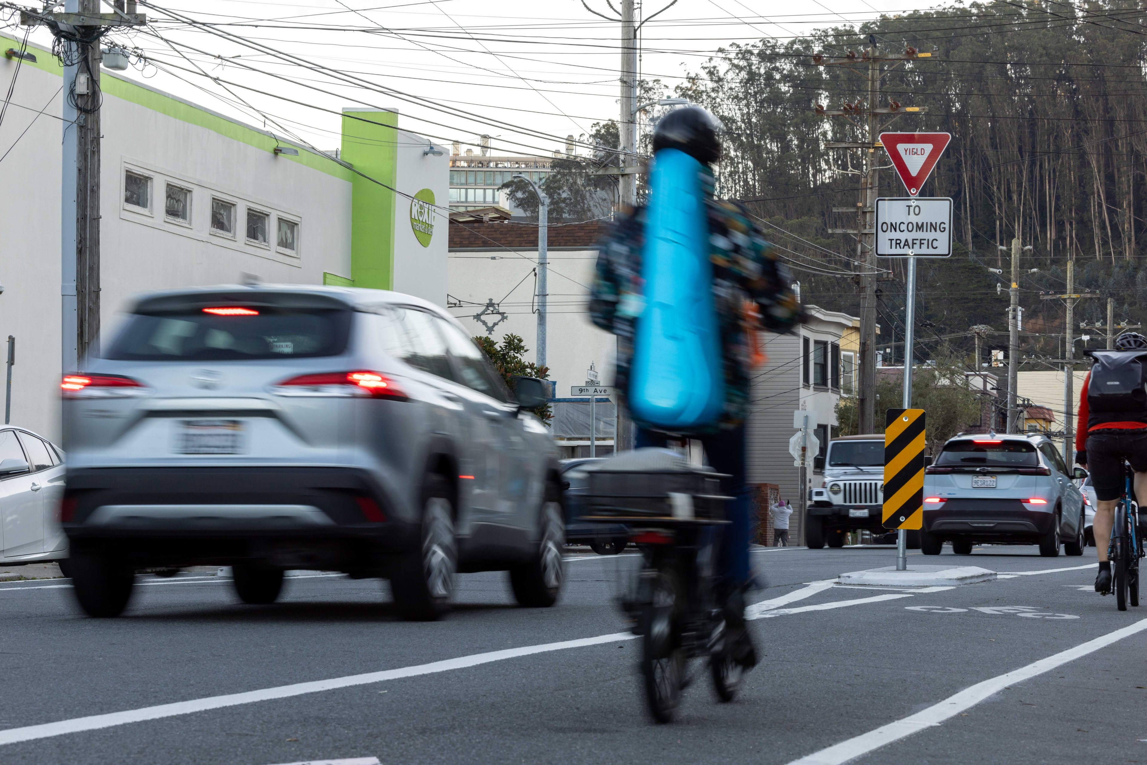 A cyclist with a blue instrument case rides alongside moving cars on a city street. A &quot;Yield to Oncoming Traffic&quot; sign is visible, with hills in the background.