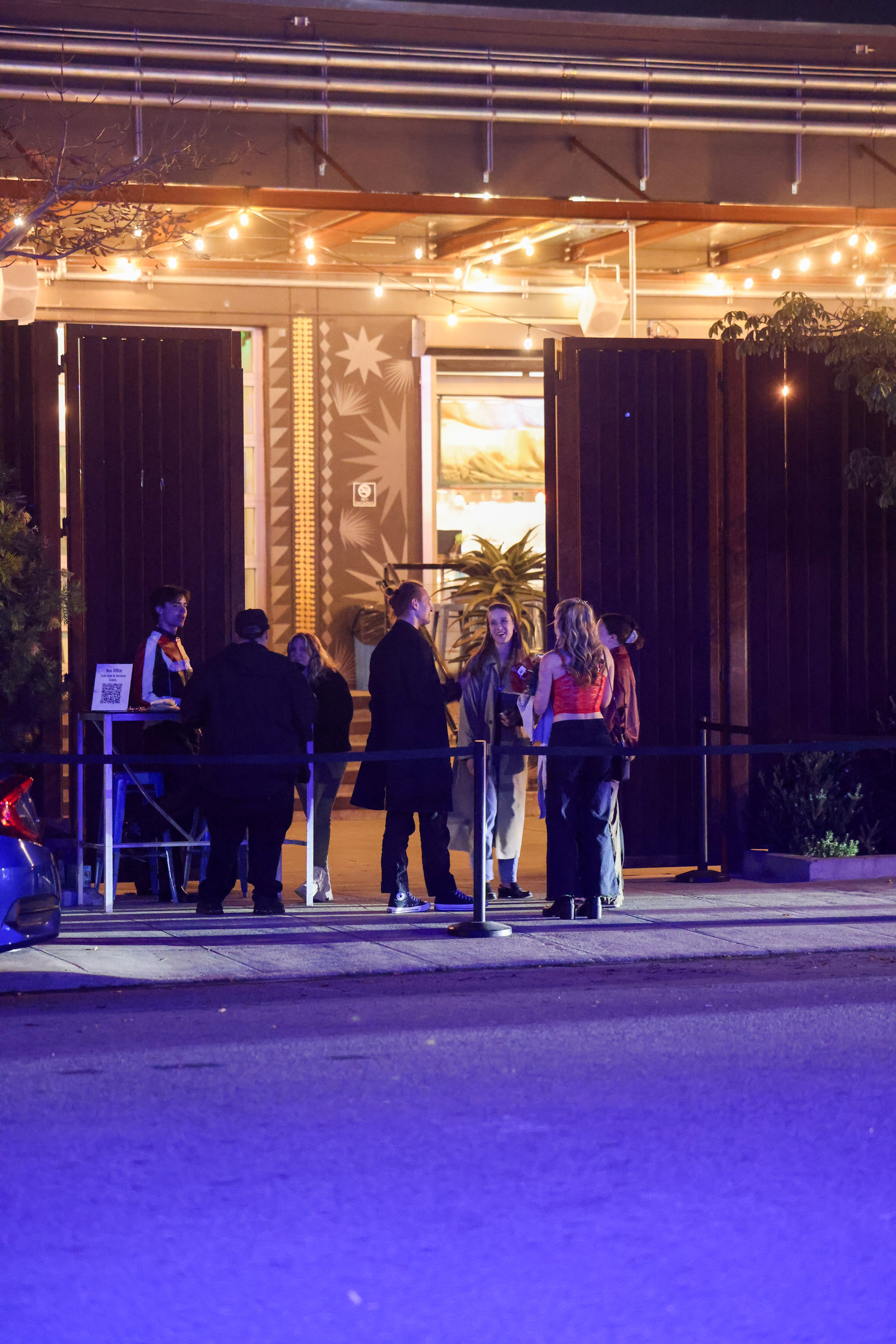 A group of people stands outside a warmly lit venue under string lights. There seems to be a check-in point with a person and a small sign.