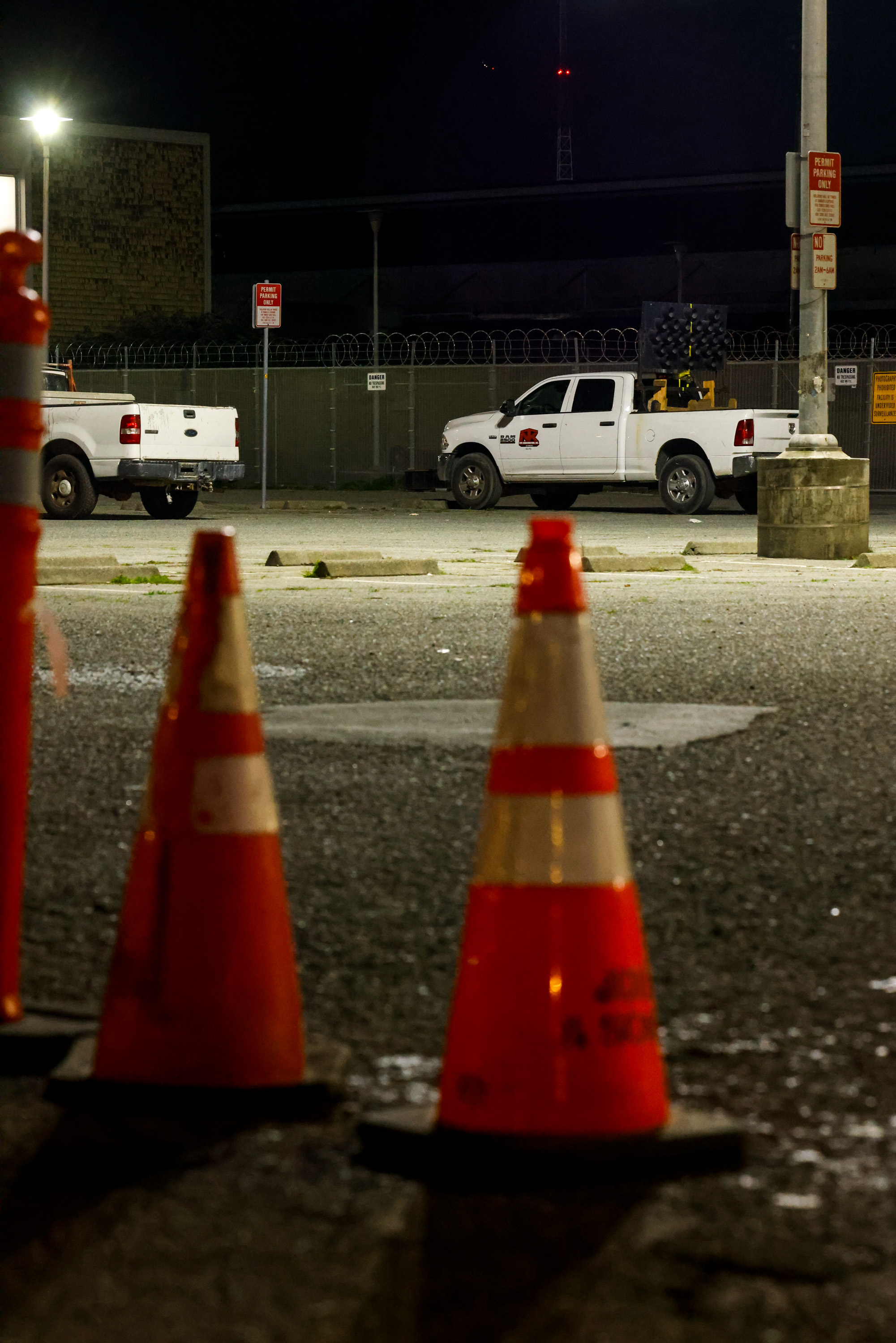 The image shows an outdoor parking lot at night with two white pickup trucks, orange cones in the foreground, and barbed wire fencing in the background.